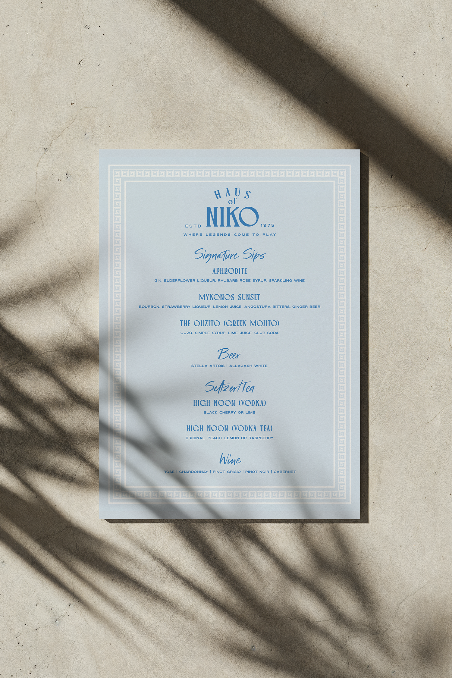A menu on a light-colored surface with shadows falling across it. The menu has a light blue background with blue text, listing various drink options from a bar or restaurant called 'Haus of Niko'.