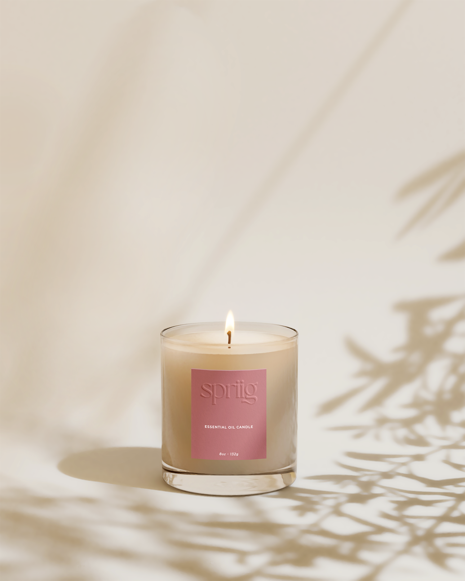 A lit essential oil candle in a clear glass container with a pink label that reads "sprig" and "essential oil candle," placed against a neutral background with shadows of foliage on the wall.