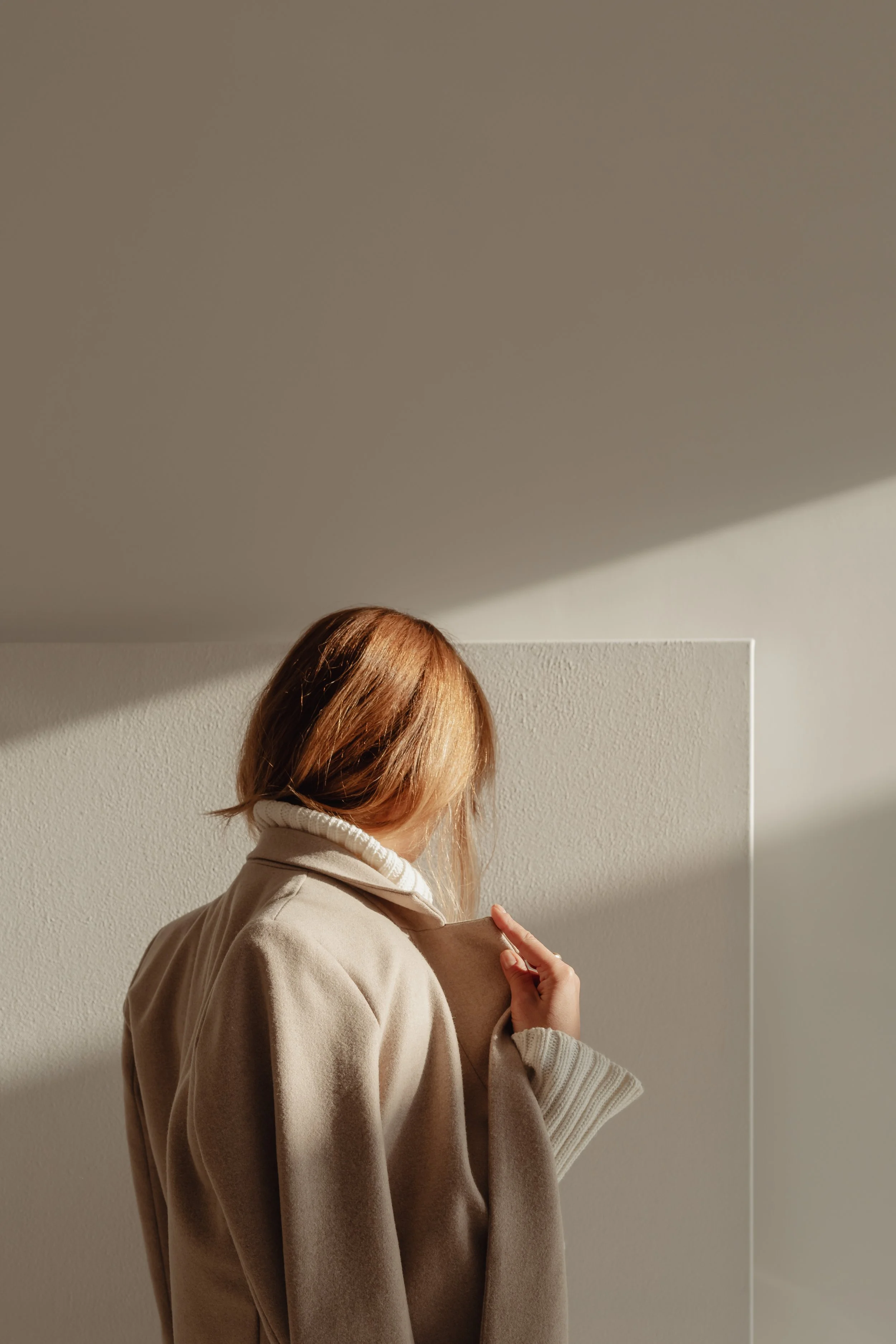 A person with reddish-brown hair, wearing a beige coat and white turtleneck, stands against a white textured wall, looking down with their hand adjusting the coat.