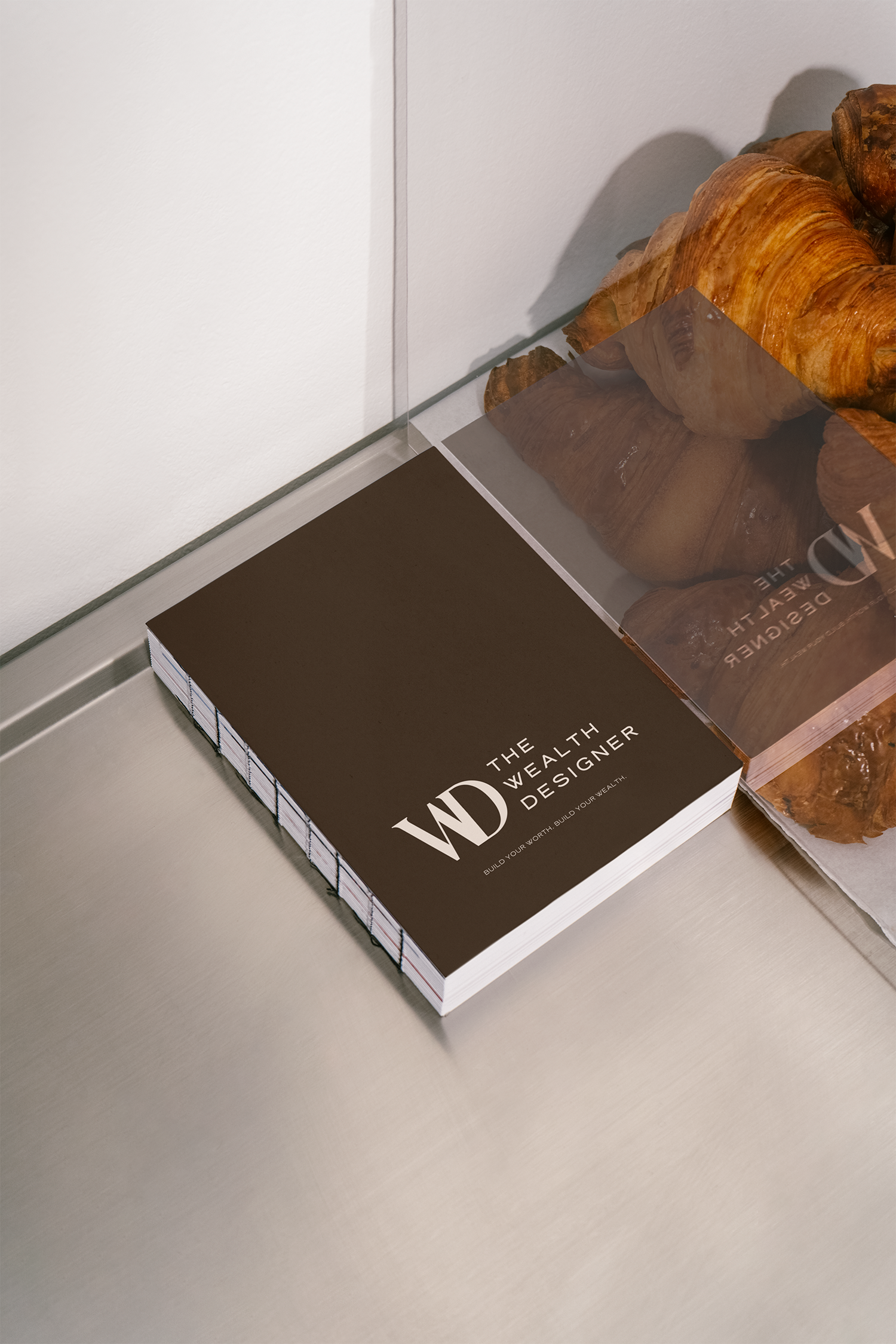 Book titled "The Wealthy Designer" on a table, with croissants on a plate in the background.