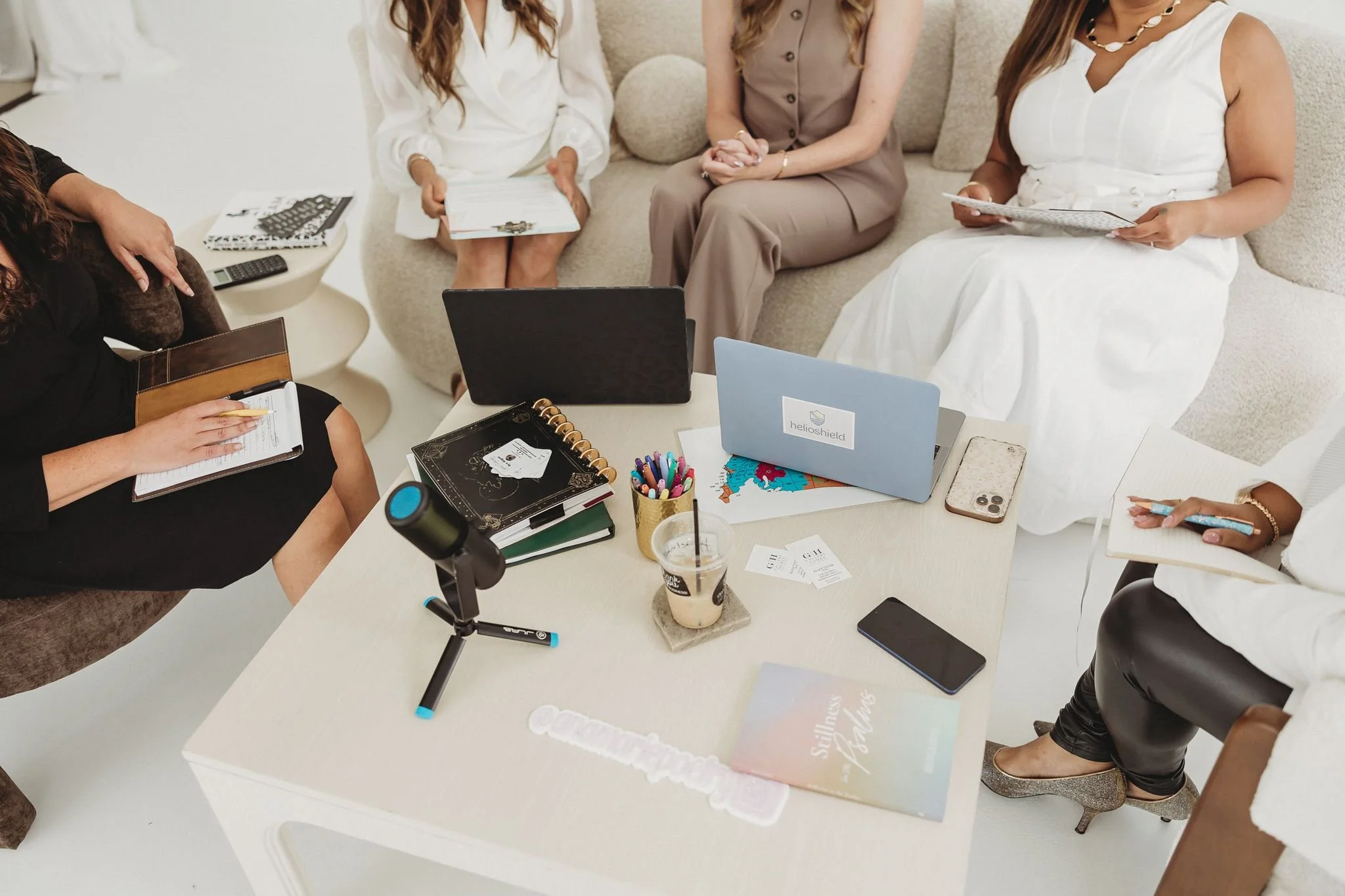 A group of women sitting around a table during a business mastermind setting, with one standing and speaking. There are coffee cups, notebooks, and glasses on the table, and a large white wall with a brick section behind them.