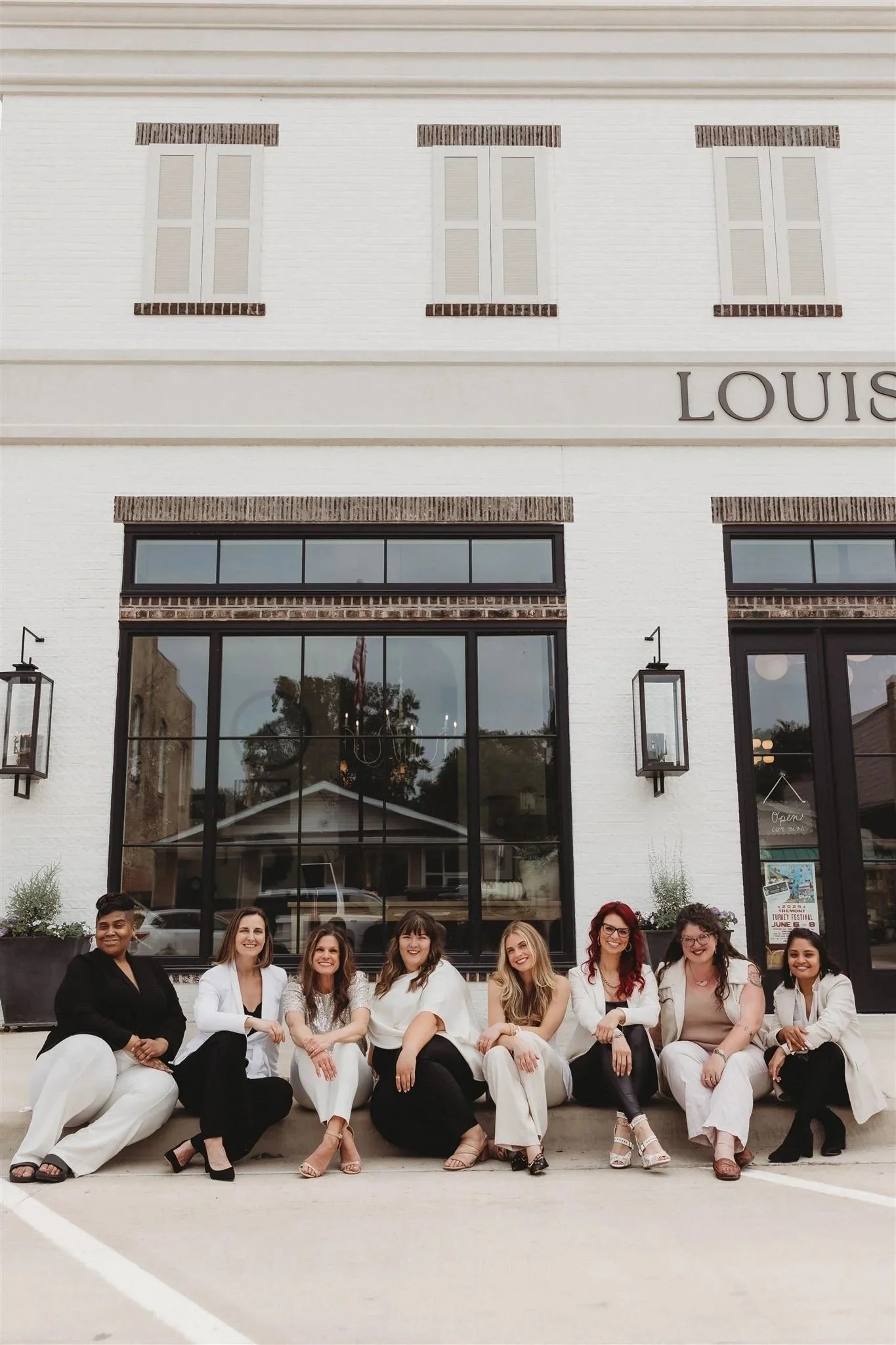 Group of women sitting on sidewalk in front of a white brick building with large windows and a sign that reads "Louis" on the wall.