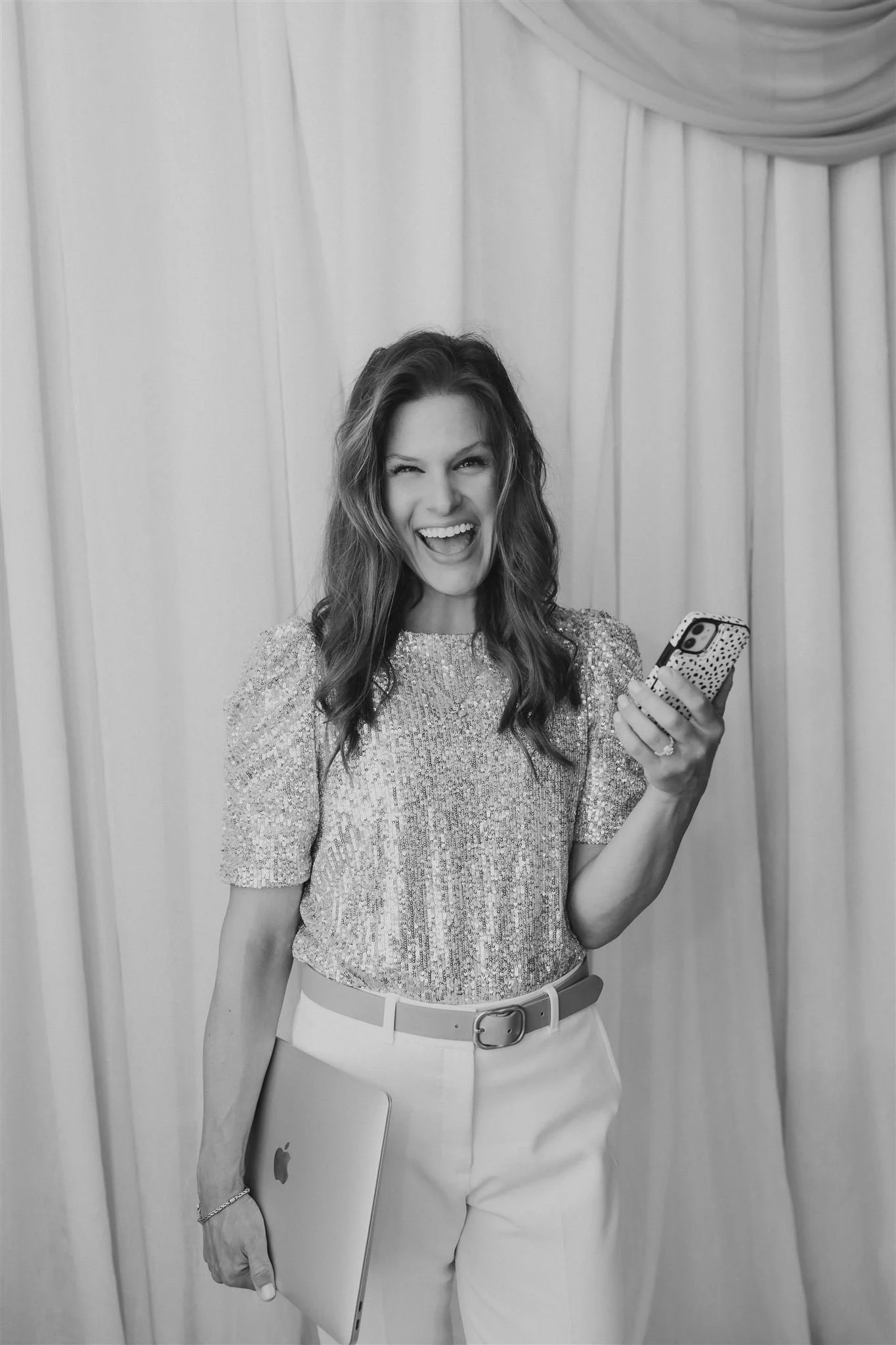 Francie Hinrichsen, founder of Founding Females, with wavy hair wearing a sparkly top, holding a laptop in one hand and a smartphone in the other, smiling in front of a curtain backdrop.