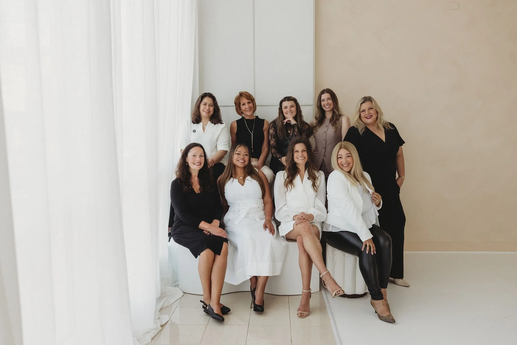 Group of nine women in elegant and professional attire, posing together indoors with a neutral-colored wall and white drapes in the background.