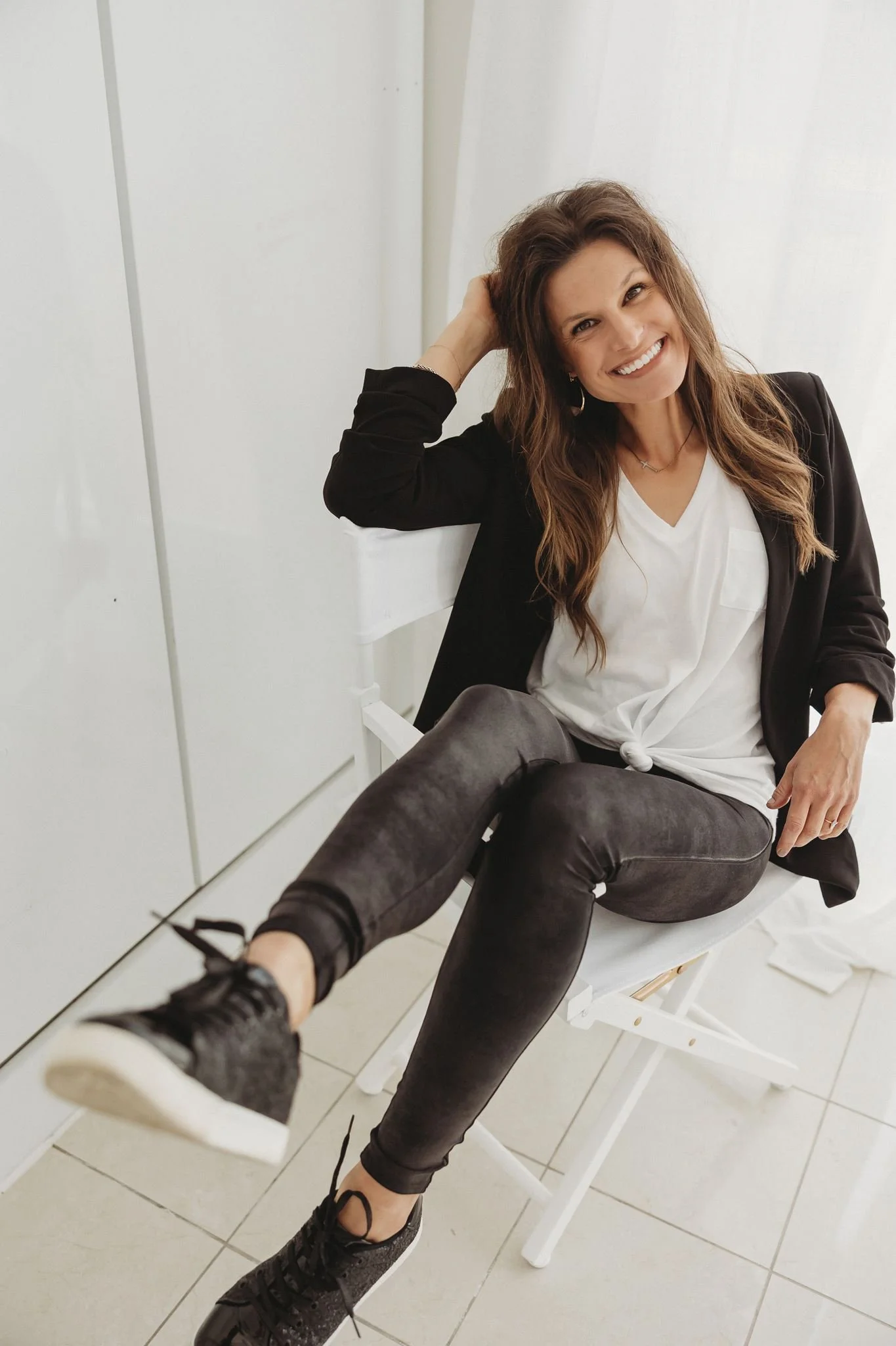A smiling Francie Mueller Hinrichsen, founder of Founding Females and SEO expert, with long brown hair sitting on a white chair, wearing a black blazer, a white top, black pants, and black sneakers, in a bright white room.