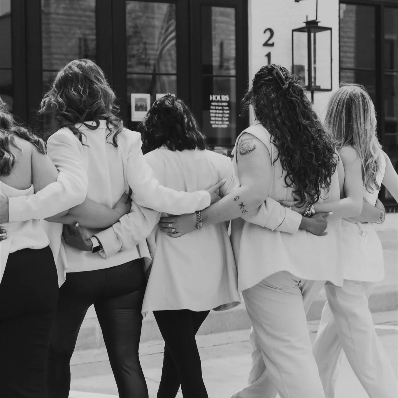 A group of women standing closely together with their arms around each other's waists, in a line, in front of a glass building entrance. The women are dressed in light-colored clothing and some have tattoos and jewelry.