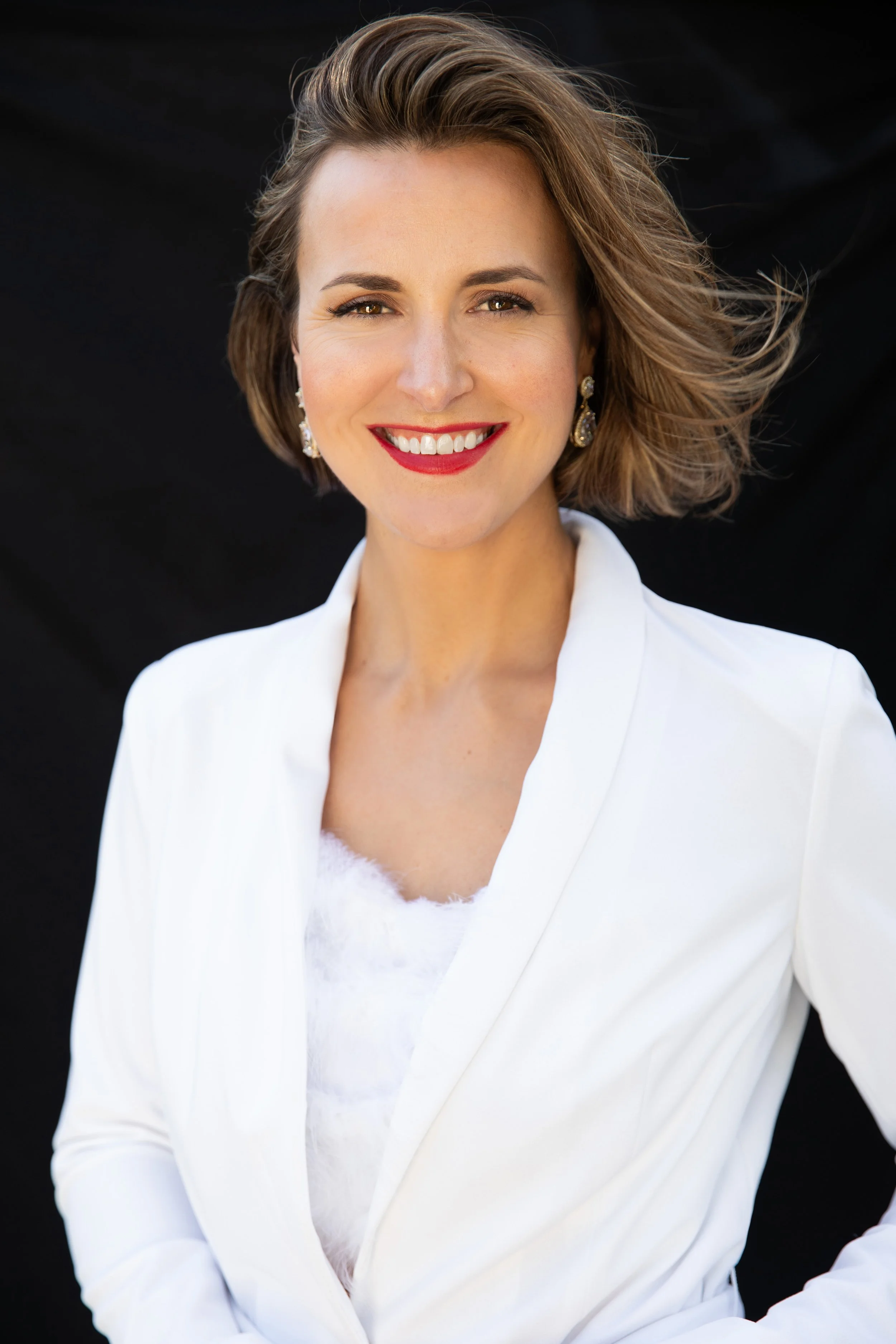 Dr. Kristen Strom, mindset and speaking coach, dressed in black clothing standing in front of a plain white wall, smiling with arms outstretched as gold confetti falls around her.