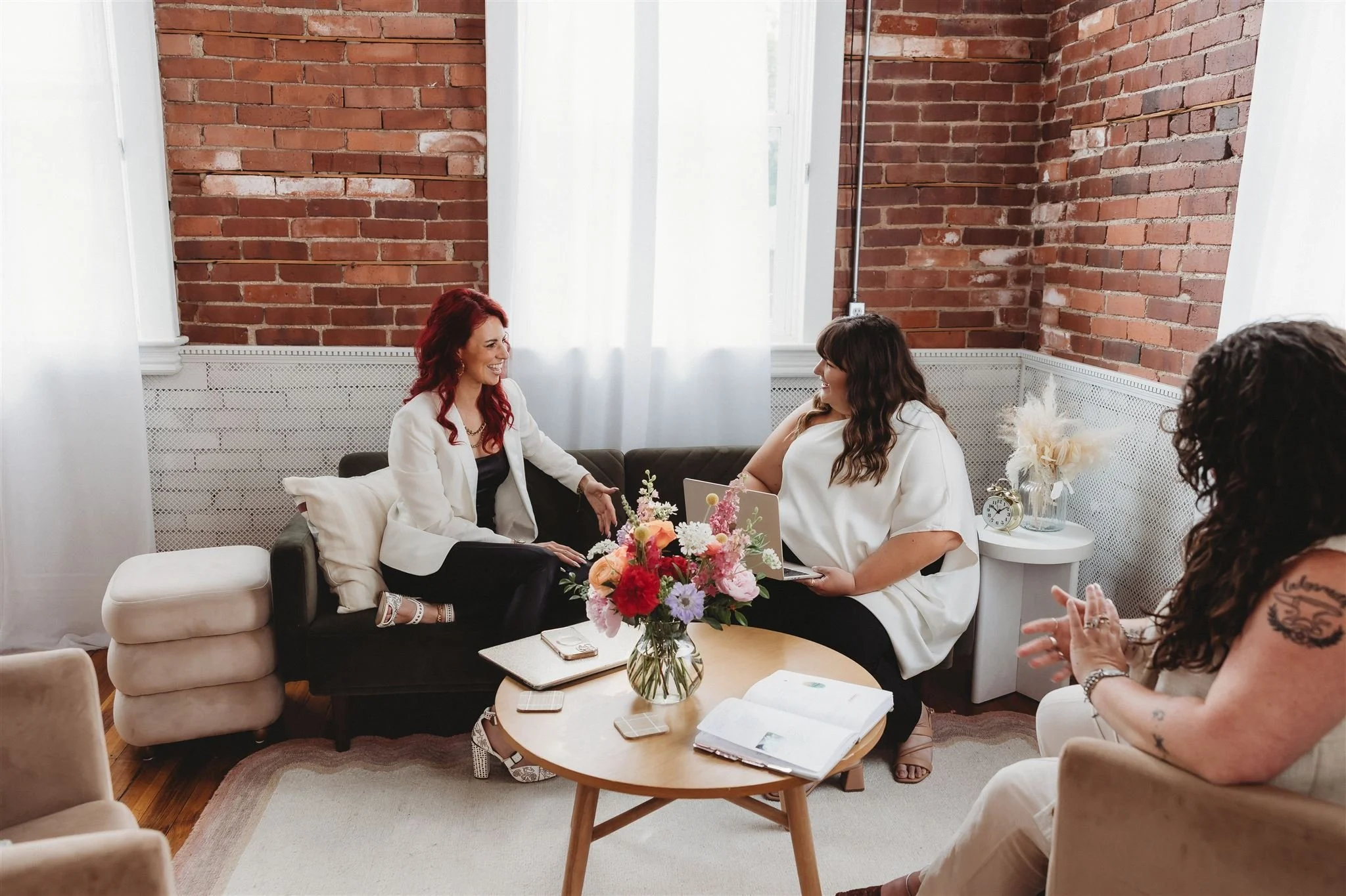 Four women sitting in a cozy room with brick walls, engaged in conversation. Two women are seated on a black sofa, smiling at each other, one holding a laptop. A woman with tattoos sits in a beige armchair, gesturing with her hands. A round wooden coffee table with a large flower arrangement, notebooks, and phones is in the center.