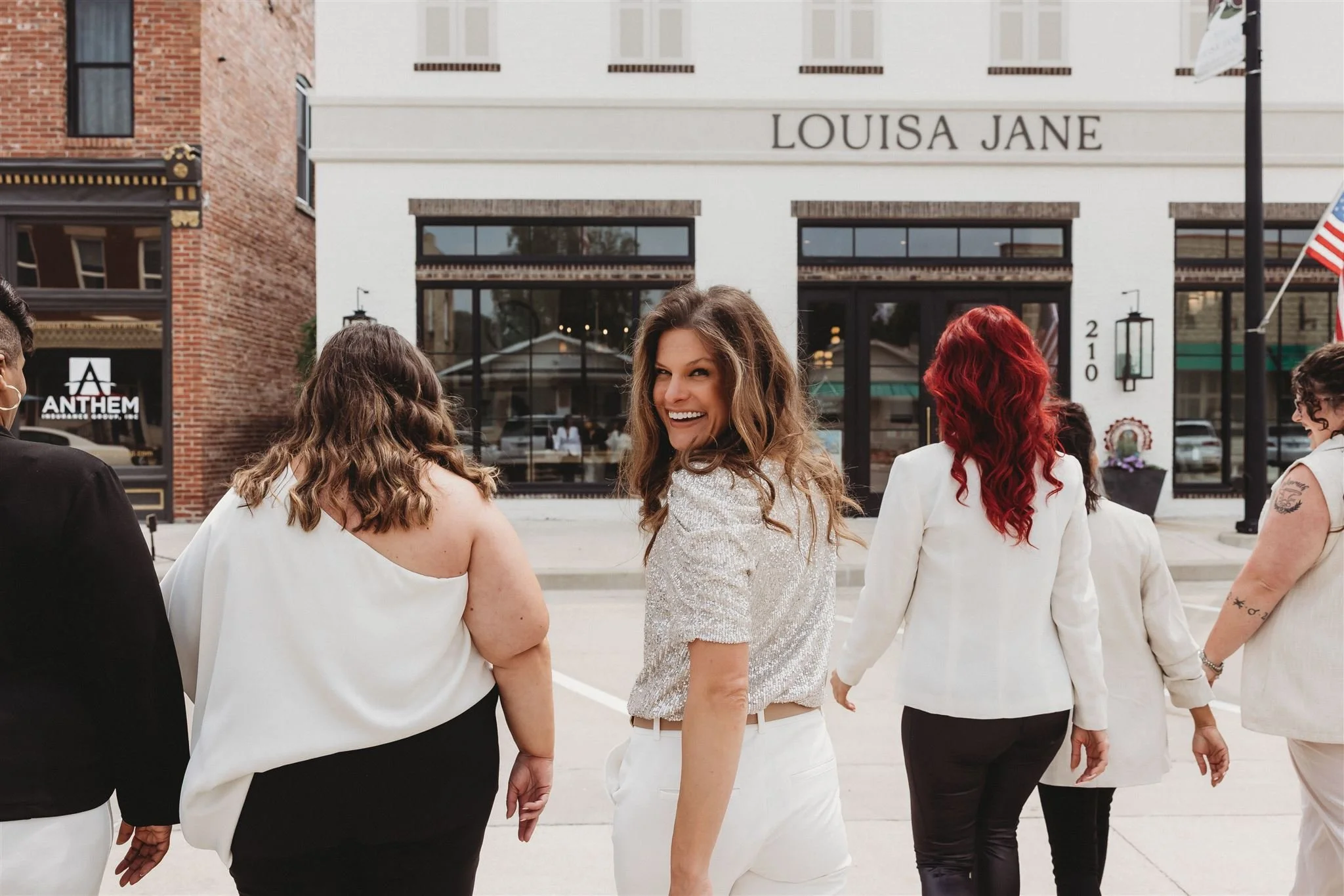 A group of diverse women business owners walking together outside in front of a locally owned design firm, "Louisa Jane." The women are dressed in white and black, with one woman smiling and looking back at the camera.
