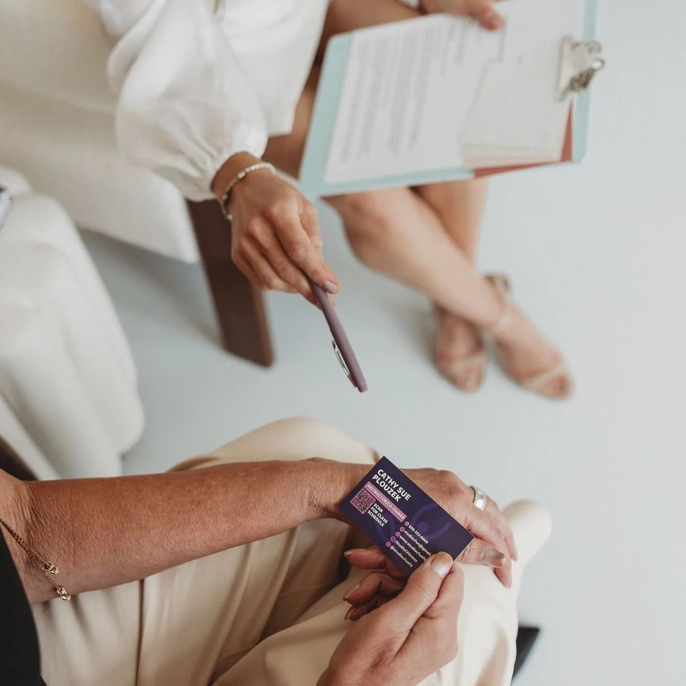 A person holding a business card, with another person sitting nearby, holding a pen and a clipboard.