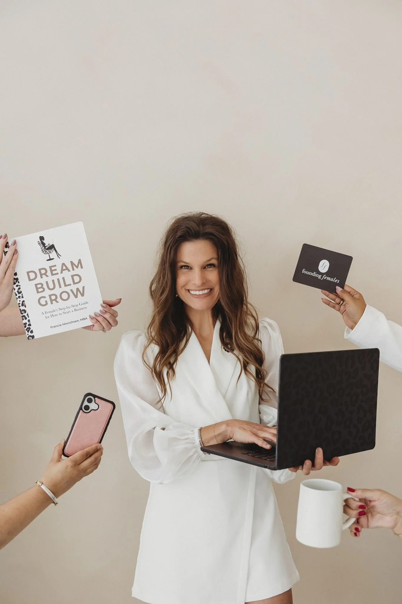 Francie Hinrichsen, founder of Founding Females, with wavy hair wearing a sparkly top, holding a laptop in one hand and a smartphone in the other, smiling in front of a curtain backdrop.