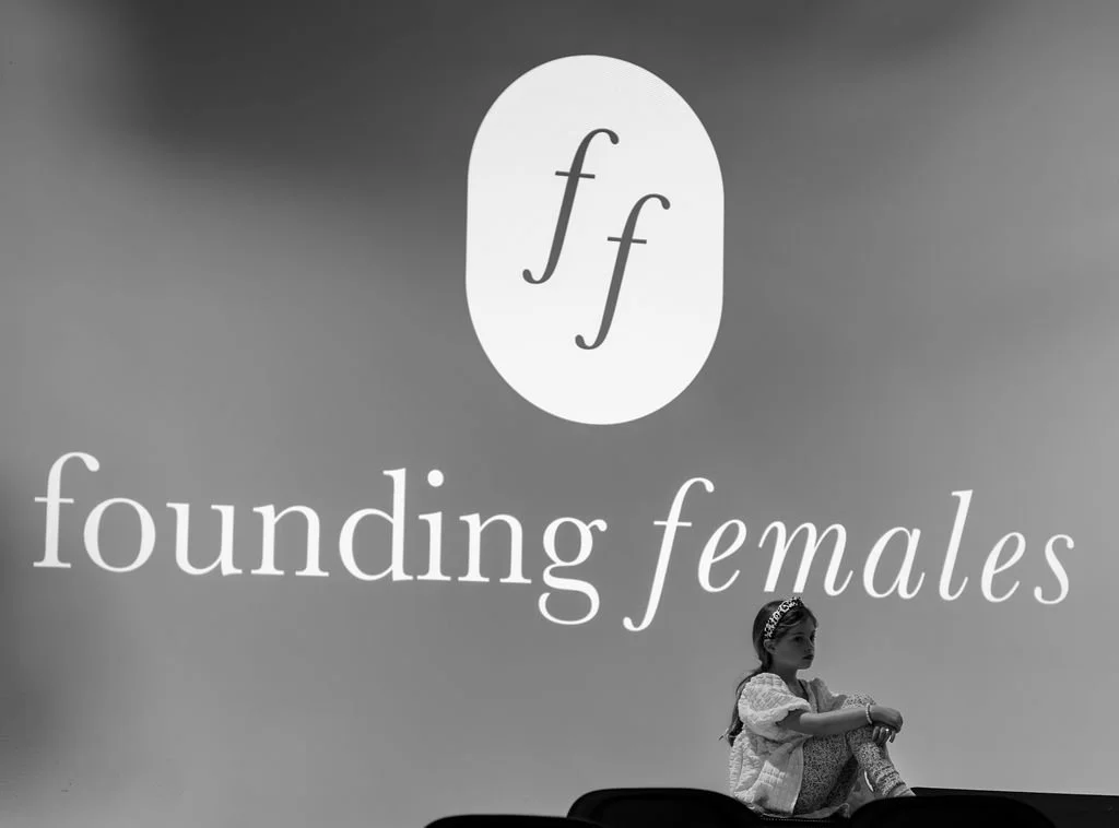 A young girl sitting cross-legged on a platform with a large sign behind her that reads 'ff founding females' and an image of two lowercase 'f's' inside an oval.
