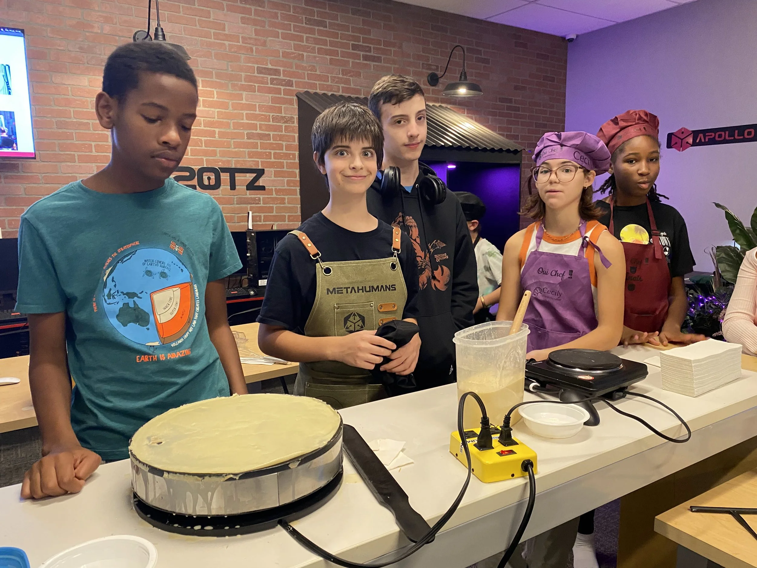 Five children standing behind a kitchen counter with cooking equipment in a room with brick walls and purple lighting, wearing aprons and chef hats, participating in a cooking activity.