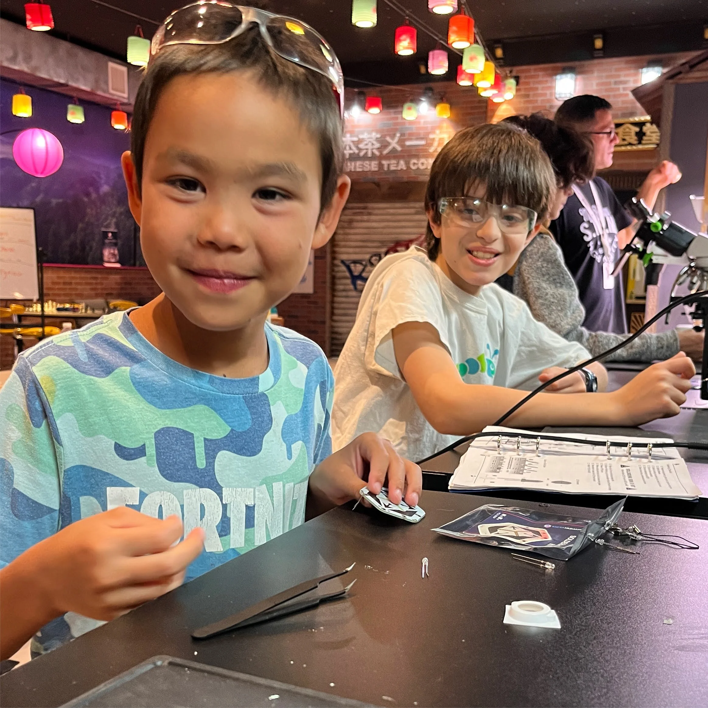 Two boys smiling at the camera, seated at a table with electronic components and tools, in a colorful indoor setting with hanging lights and a brick wall in the background.