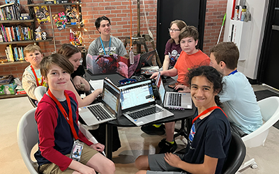 A group of children and teenagers working on laptops around a round table in a classroom or library setting.