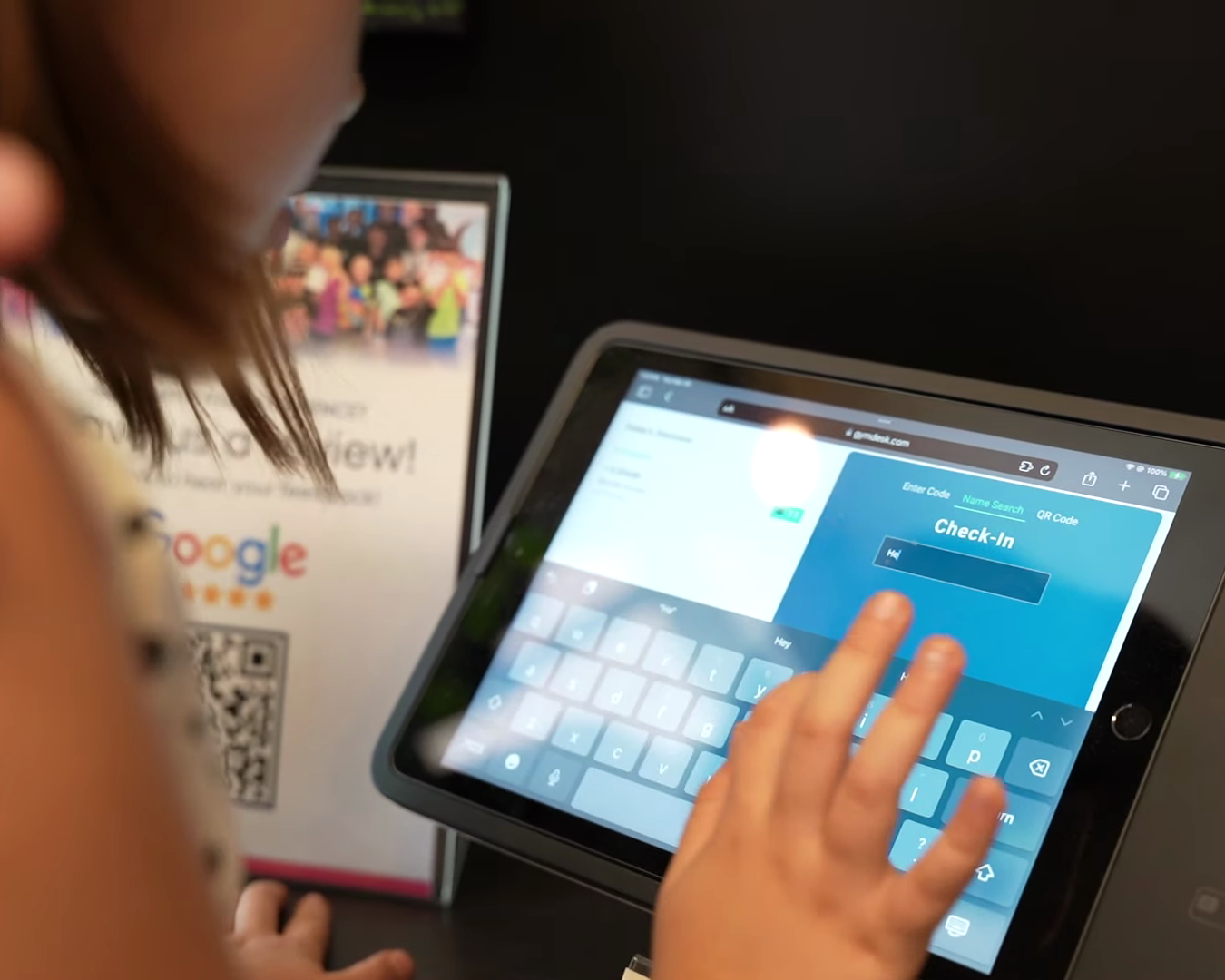 A person using a tablet with a check-in screen on the display, tapping the screen. In the background, there is a blurred sign with the Google logo and a QR code.
