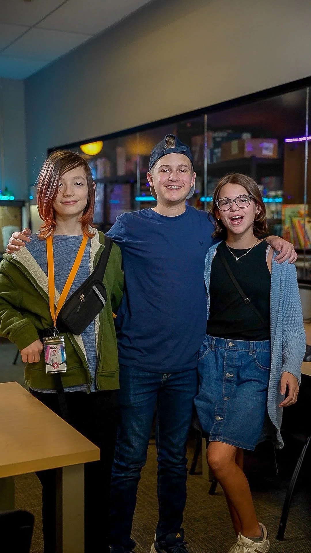 Three teenagers standing together in a room, smiling at the camera. Two girls and one boy, with arms around each other, in a setting with books and shelves in the background.