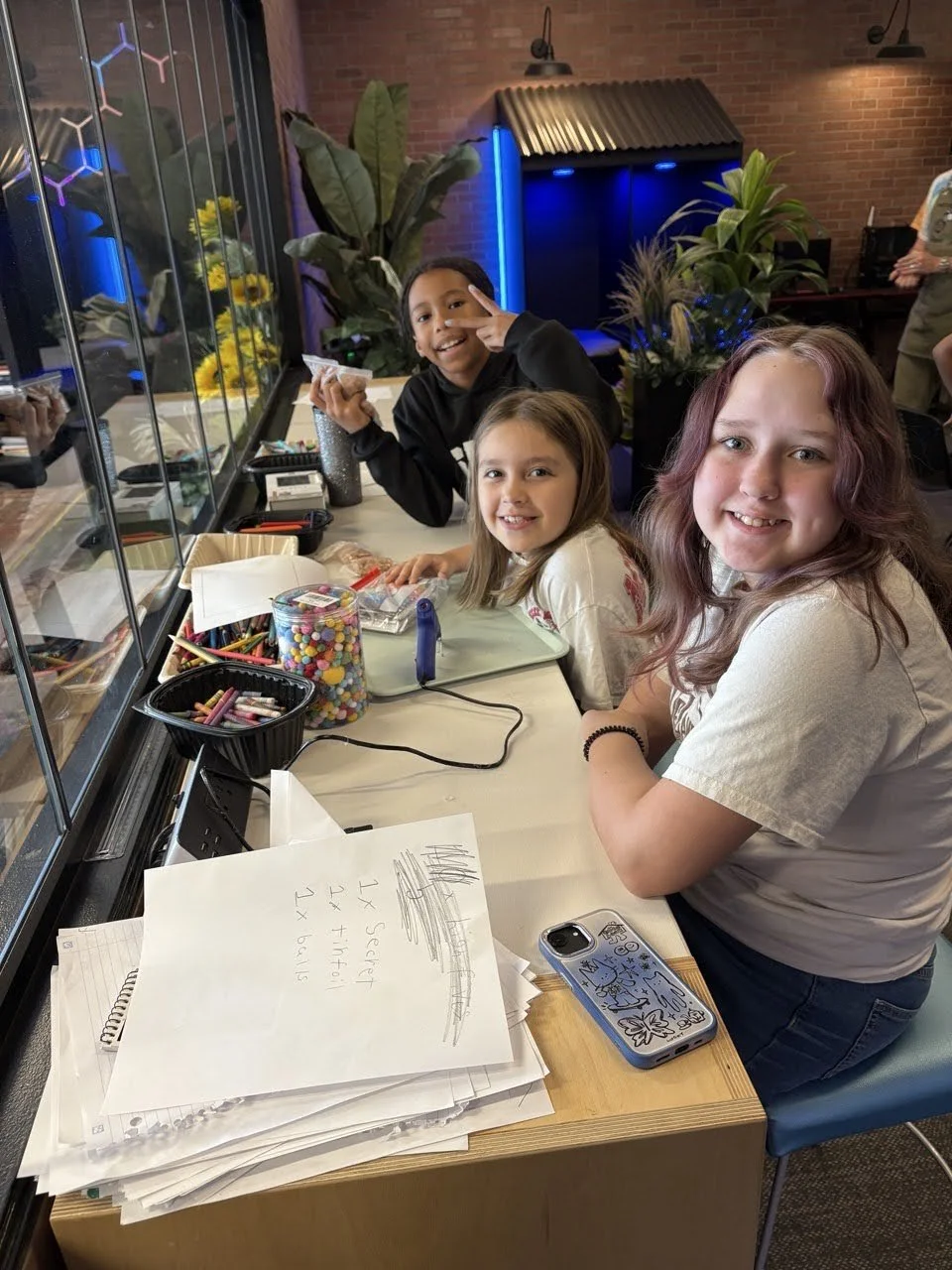 Three girls sitting at a table covered with art supplies, smiling and looking at the camera, in a room with plants and brick walls.