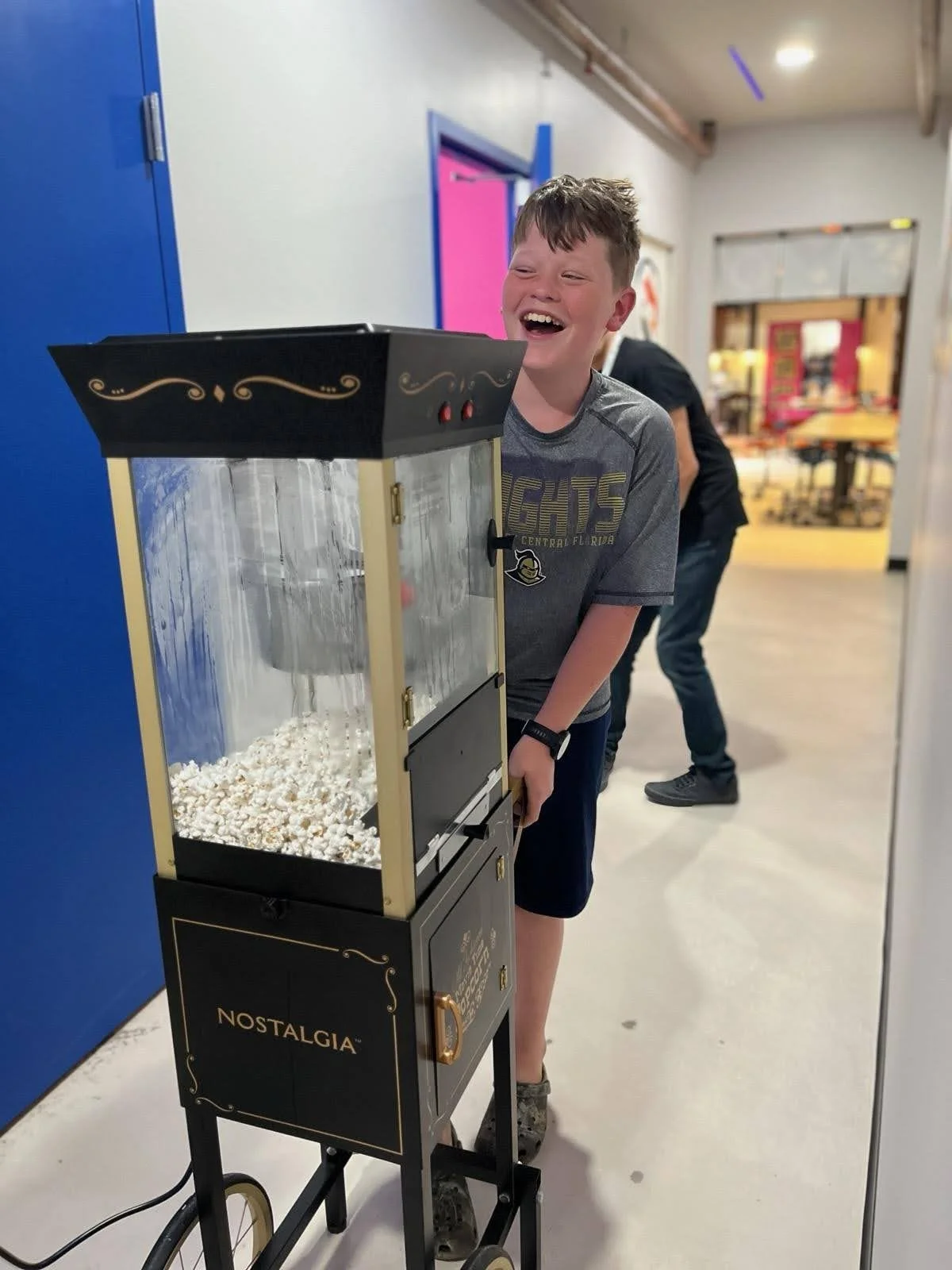 A boy laughing while standing behind a popcorn machine labeled 'Nostalgia' in a colorful indoor setting.