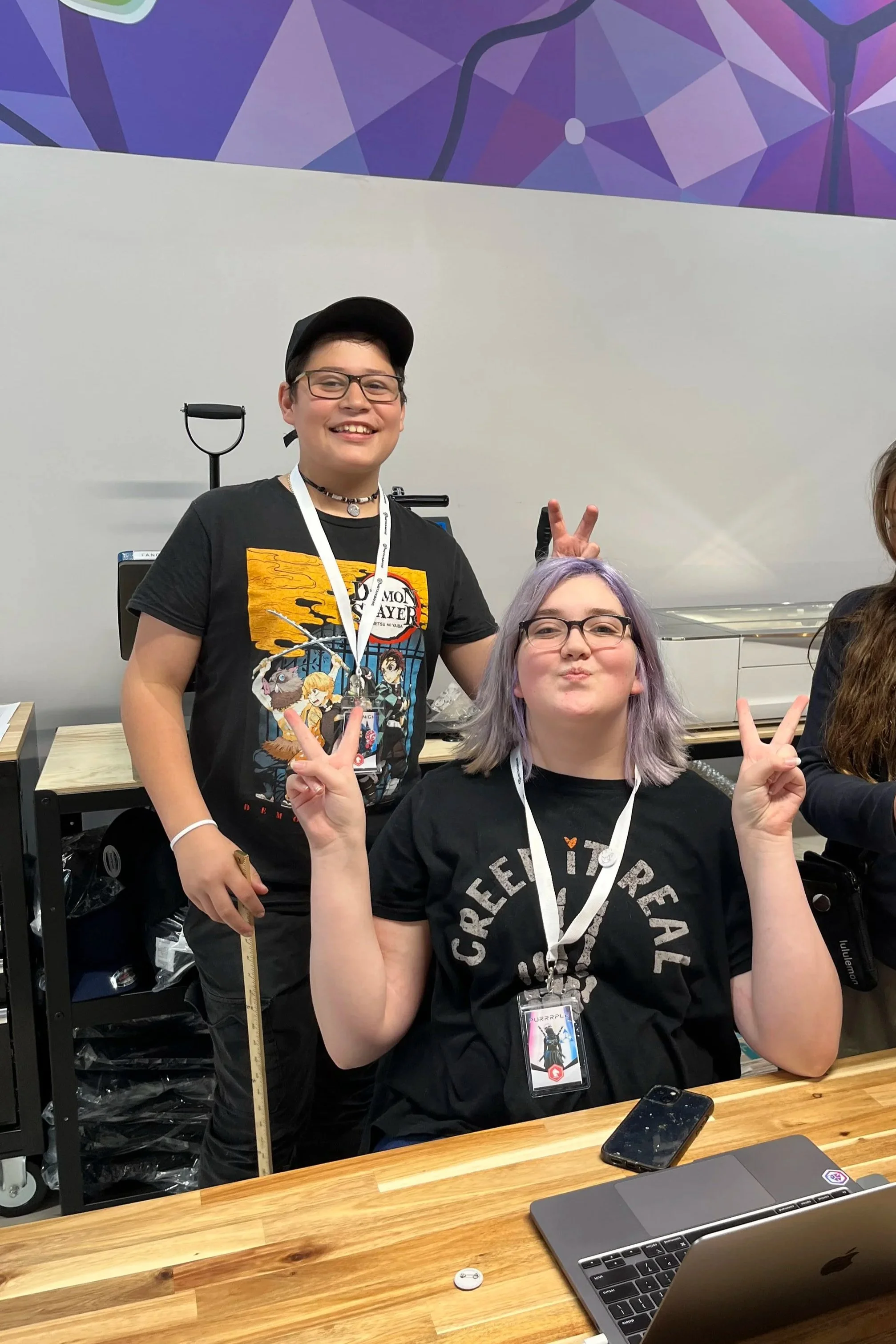 Two young women at a table, one standing and one sitting, making peace signs and smiling, with a purple geometric mural in the background.