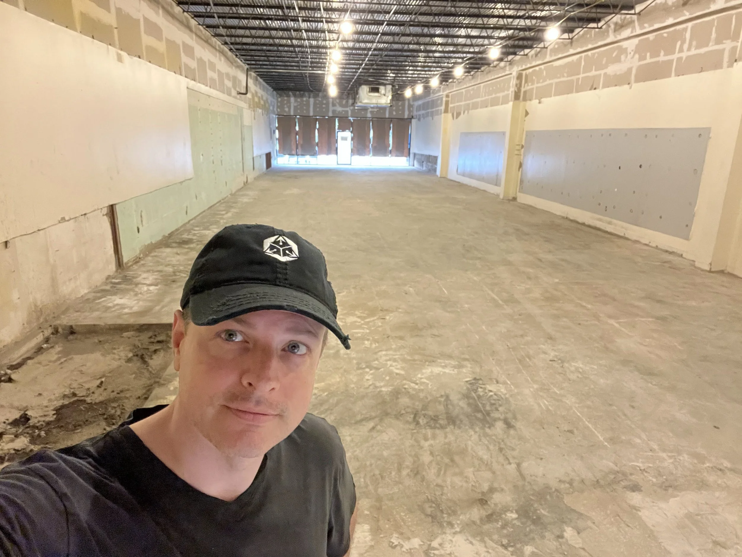 A man wearing a black cap and black T-shirt taking a selfie inside an unfinished commercial space with exposed walls, ceiling, and concrete floor.