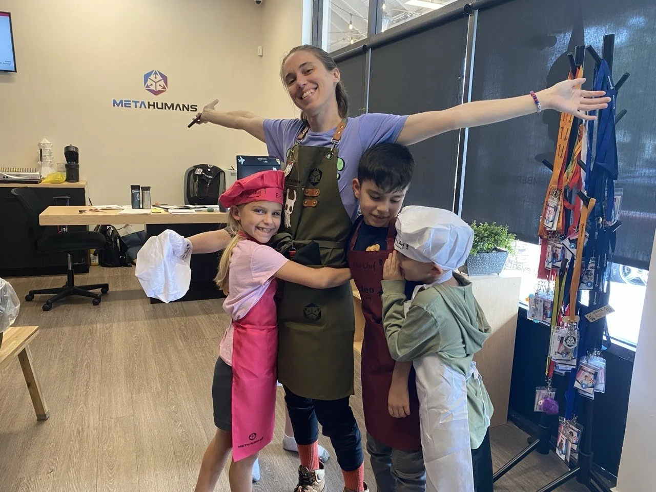 Smiling woman with arms outstretched, surrounded by three children dressed as chefs, in a room with a METAHUMANS logo on the wall and kitchen utensils, in a fun, playful setting.
