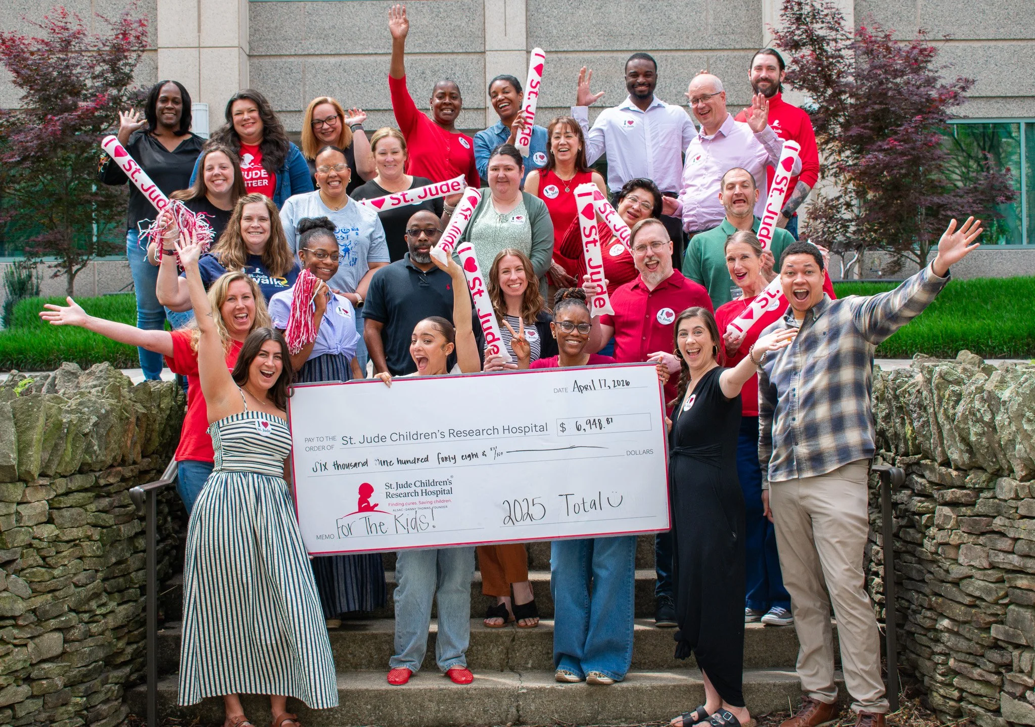 Group of diverse people celebrating outdoors, holding a large donation check for $6,948.87 to St. Jude Children's Research Hospital, with some waving and smiling.