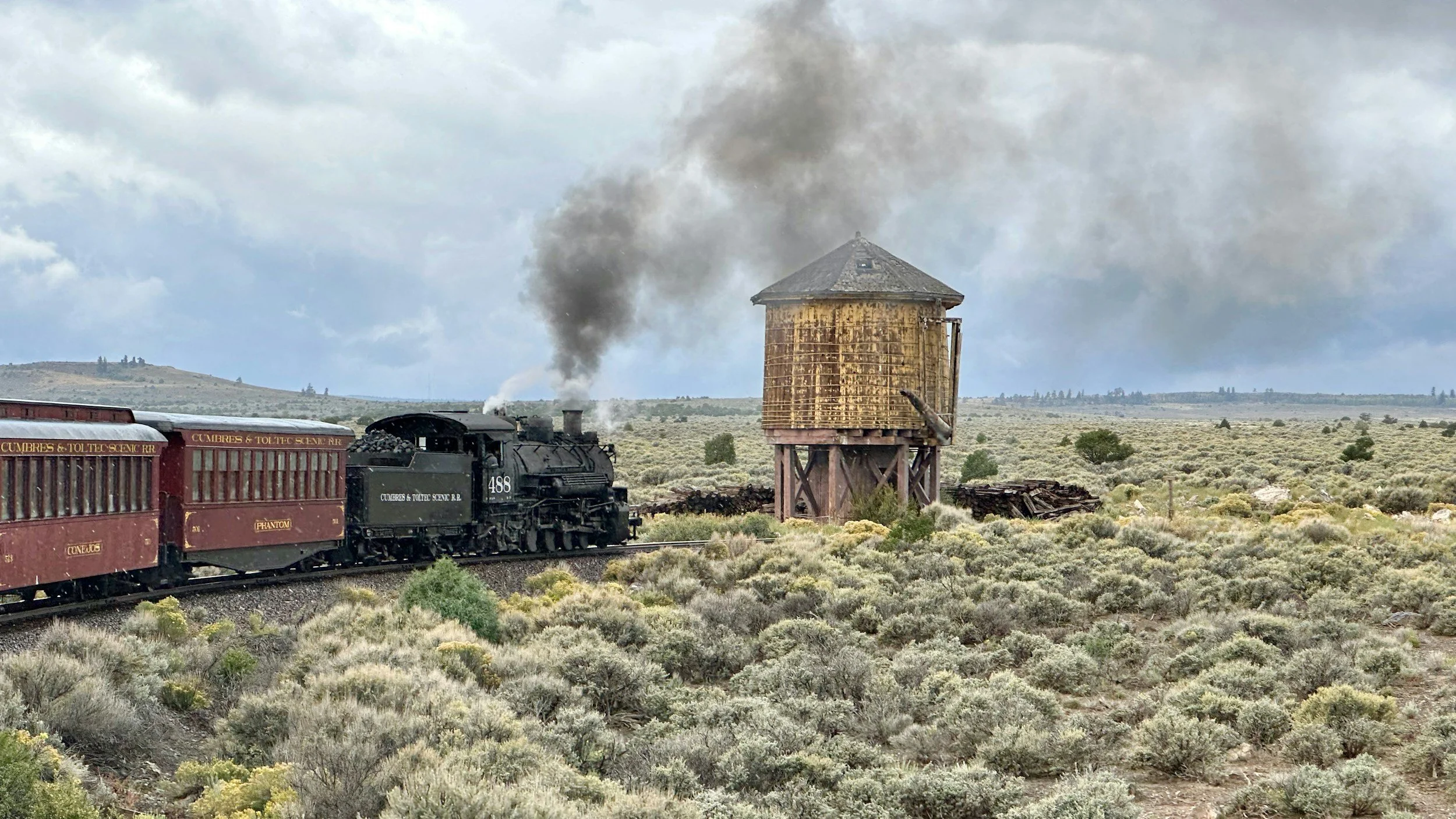 A black steam locomotive pulling red passenger cars labeled "Cumbres & Toltec Scenic RR" travels through a desert landscape, with an old wooden water tower emitting dark smoke in the background, under a cloudy sky.