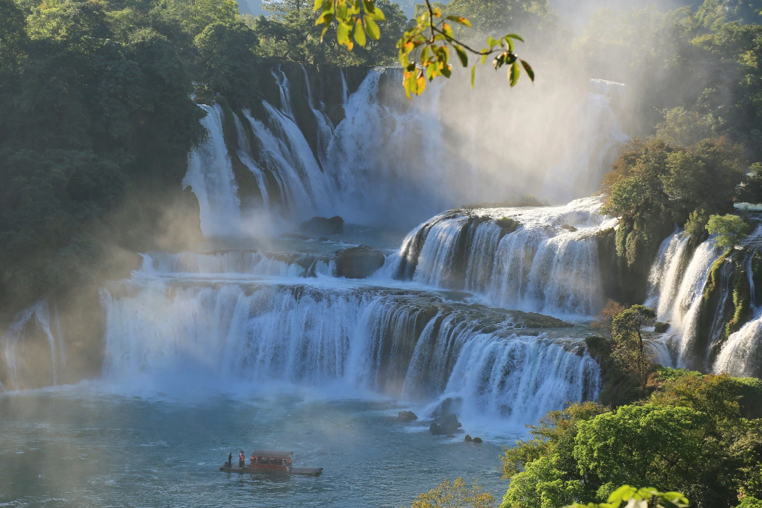 Scenic waterfall with multiple tiers of cascading water surrounded by lush green trees, and a boat with people near the base.