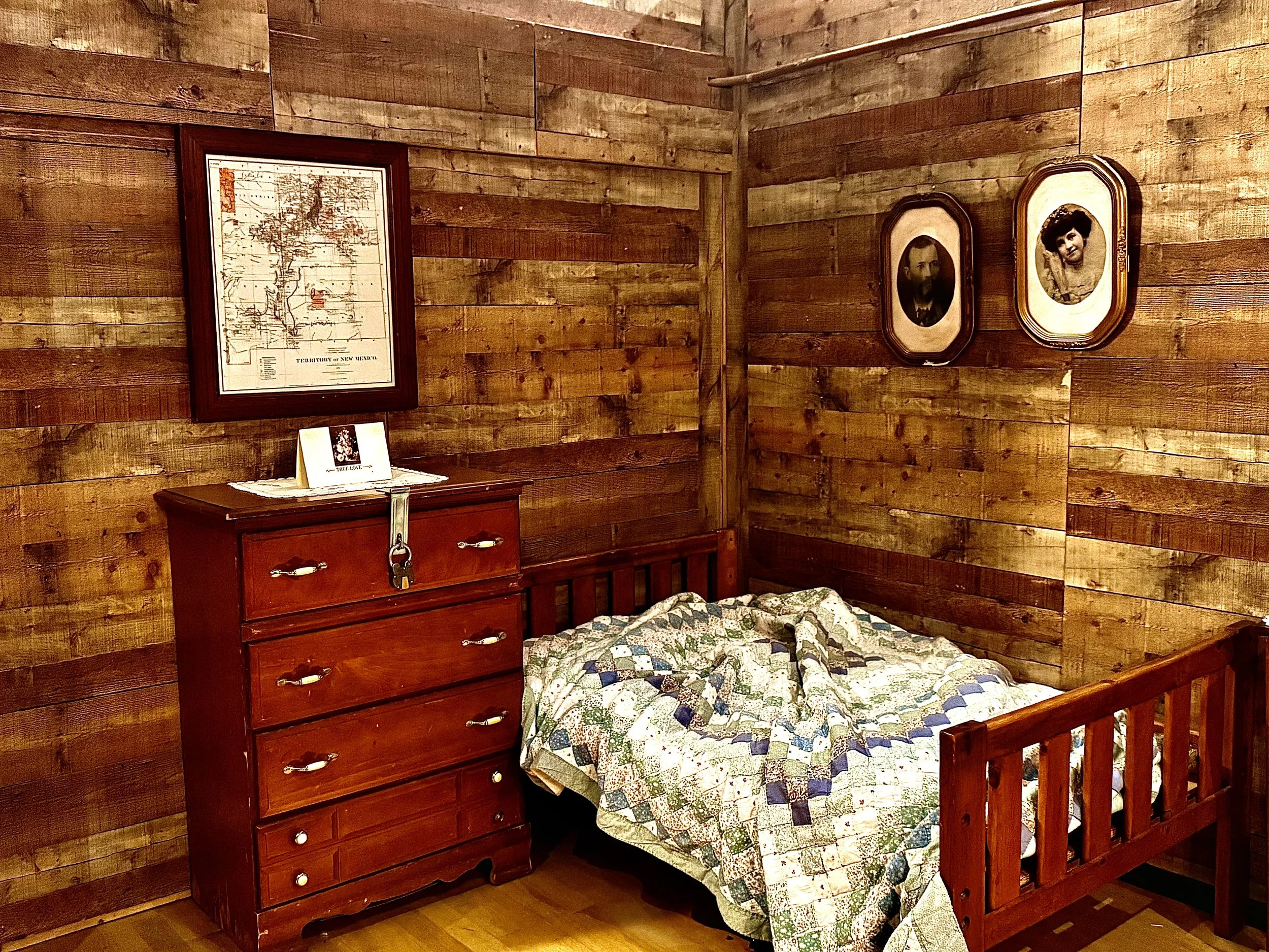 A rustic bedroom with wooden plank walls, a red wooden dresser with a lock, a bed with a colorful quilt, and framed photos on the wall.