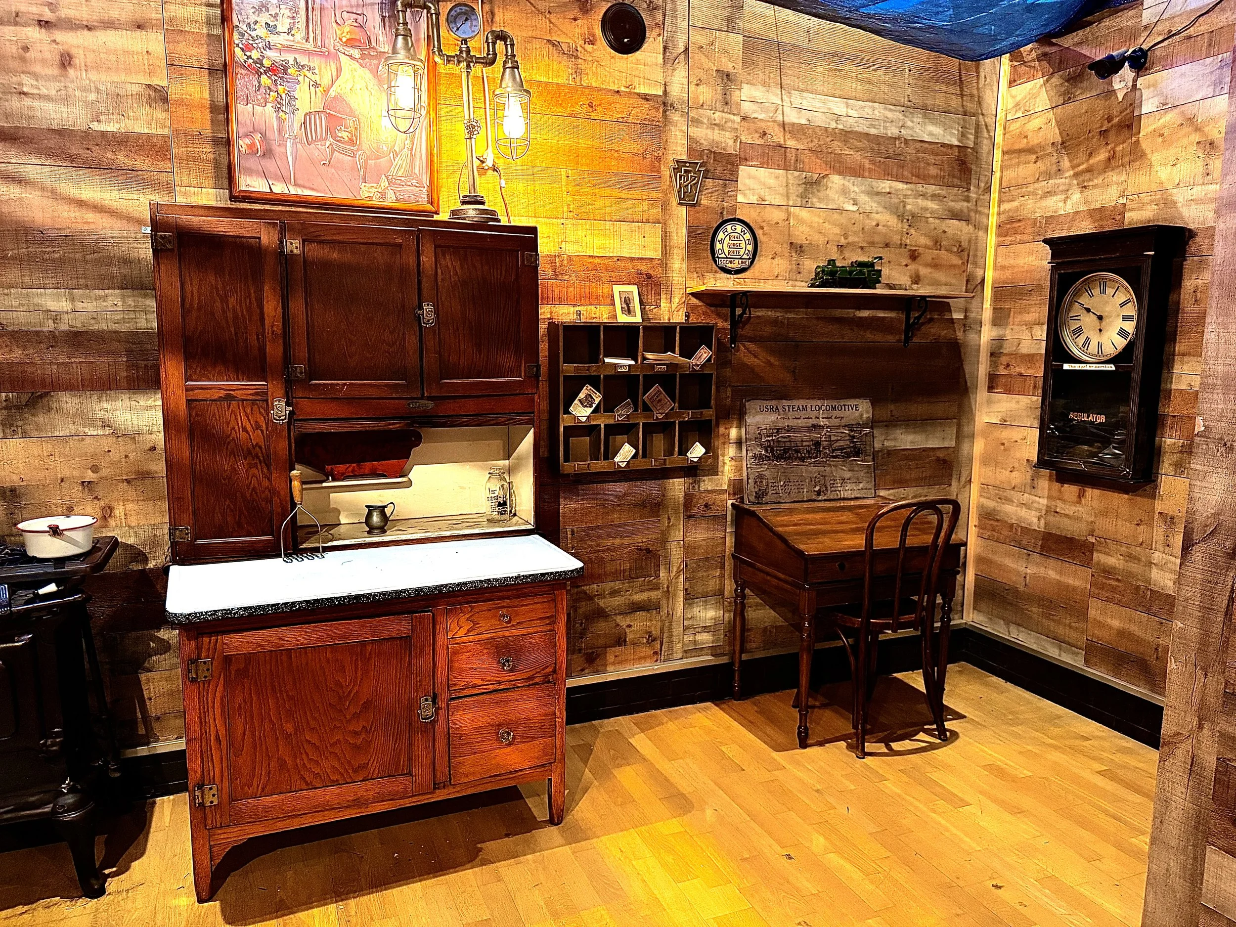 A cozy vintage-style room with wooden walls and floor, featuring a wooden cabinet, a small table with a single chair, wall clocks, and various decorative items.