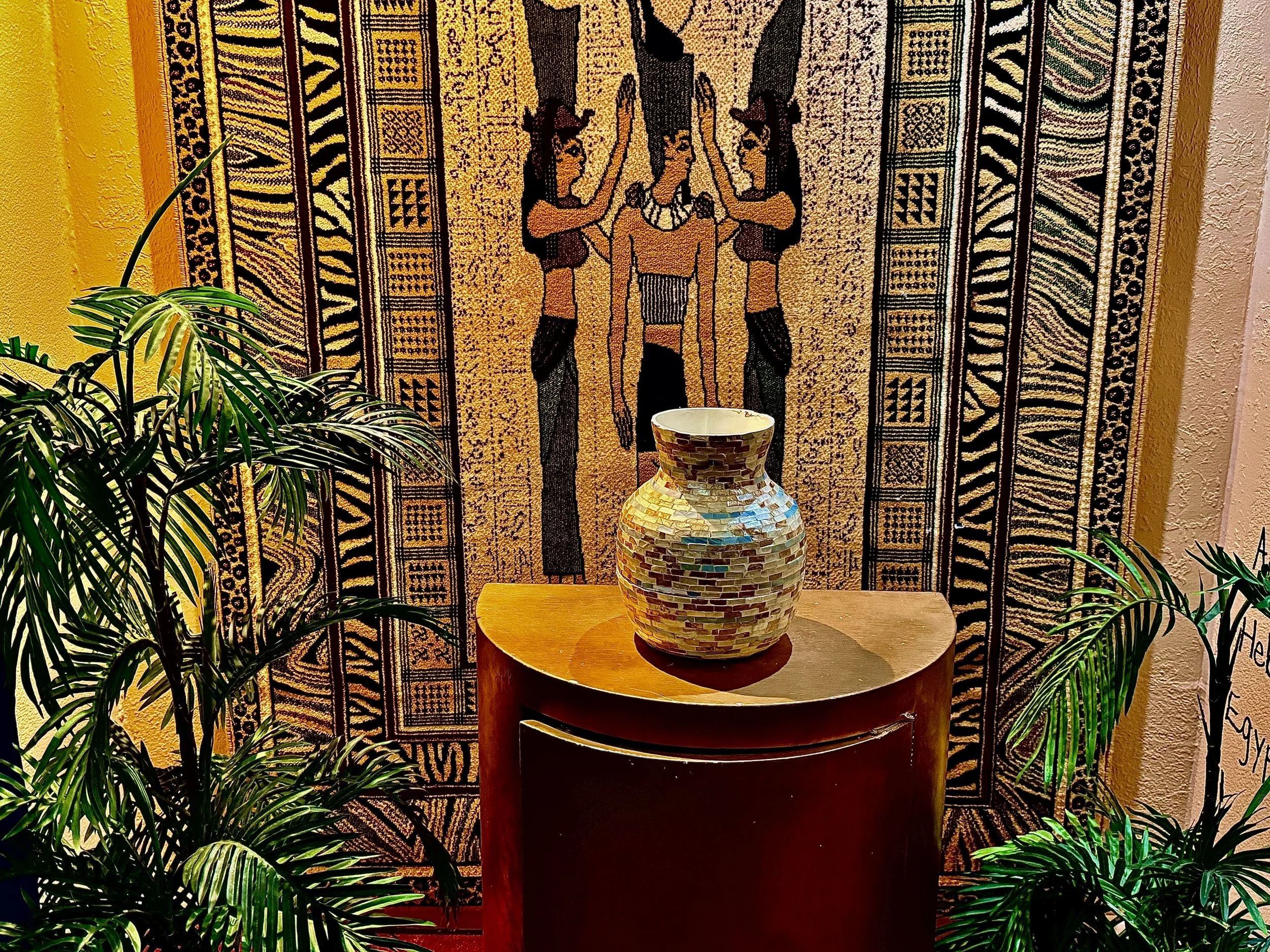 A ceramic vase with multicolored mosaic pattern on a wooden stand, in front of a traditional African textile wall hanging, surrounded by green palm plants.