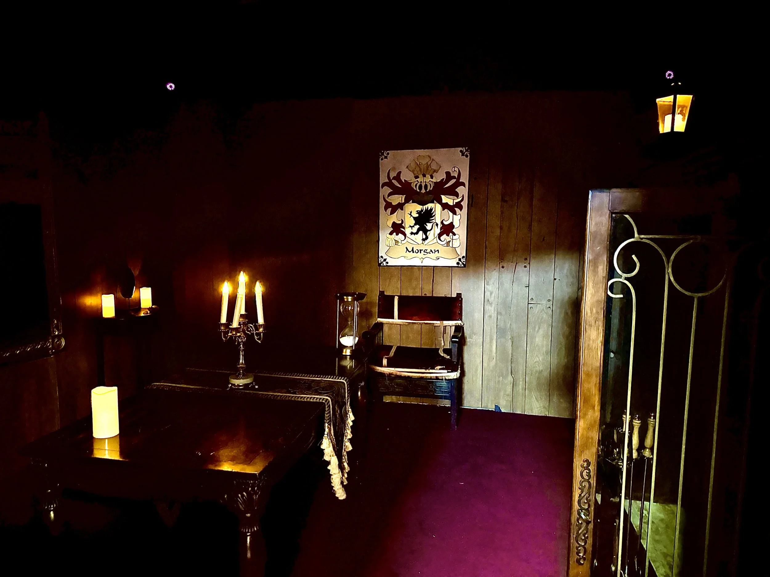 A dimly lit room with wooden walls and red carpet, decorated with candles and a framed heraldic shield on the wall labeled 'Morsan'.
