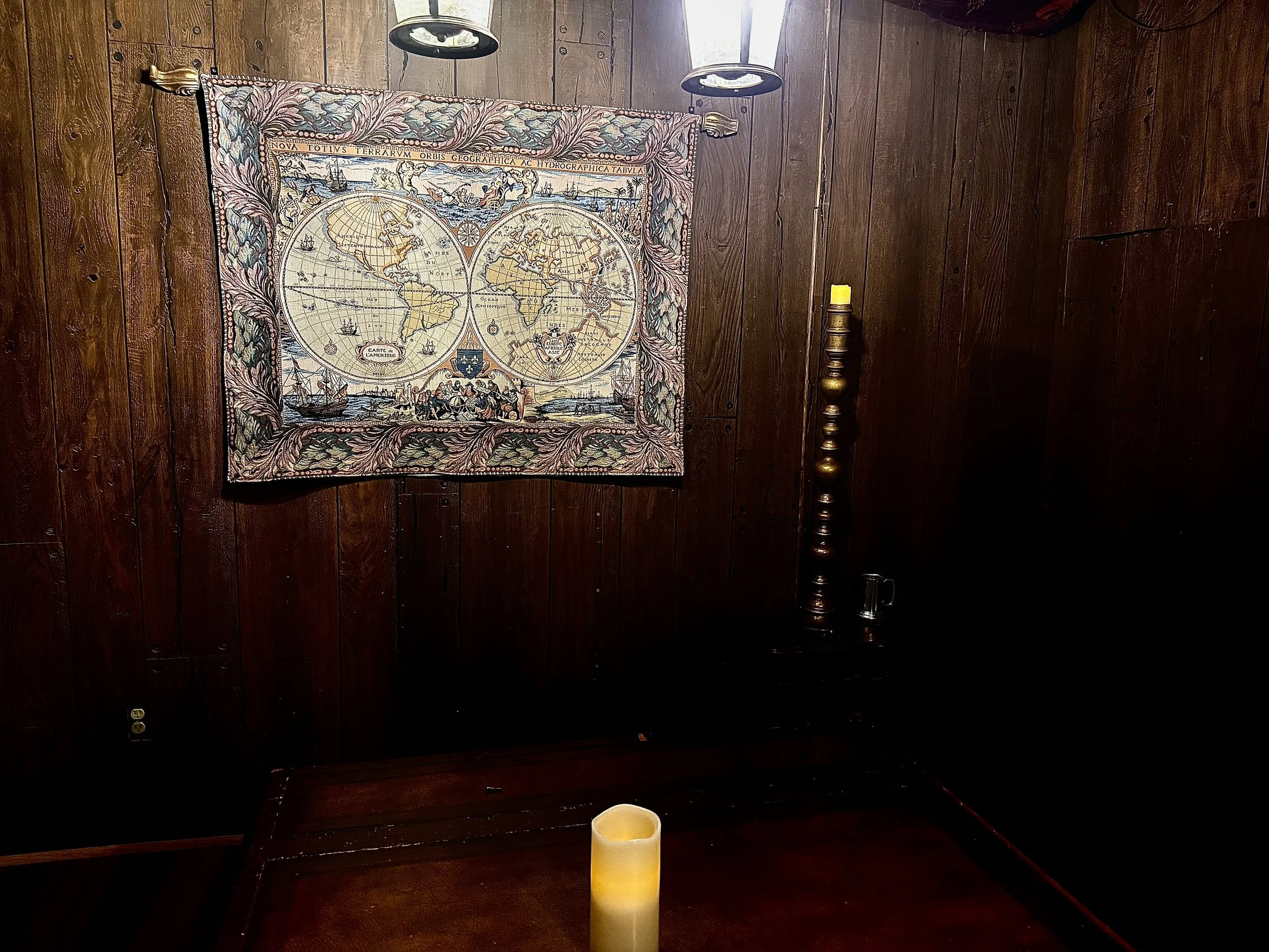 A dark wood-paneled room with a decorative antique world map tapestry on the wall, a yellow candle on the table, a tall candle holder with a yellow candle, and a small glass mug on a side table.