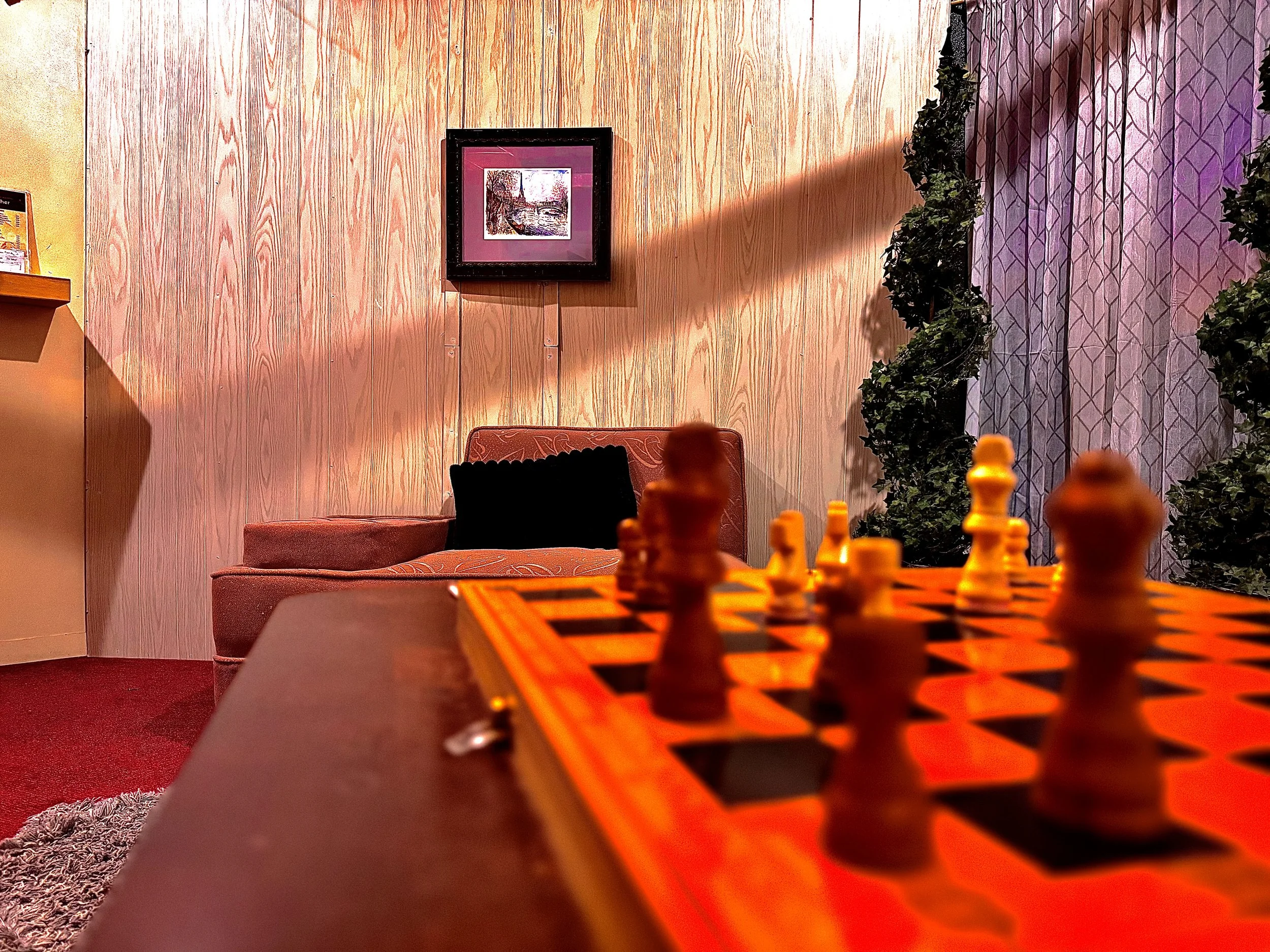 A chessboard with chess pieces set up on a table in the foreground, a sofa with a black pillow, a framed picture on a wooden wall, and greenery with curtains in the background.