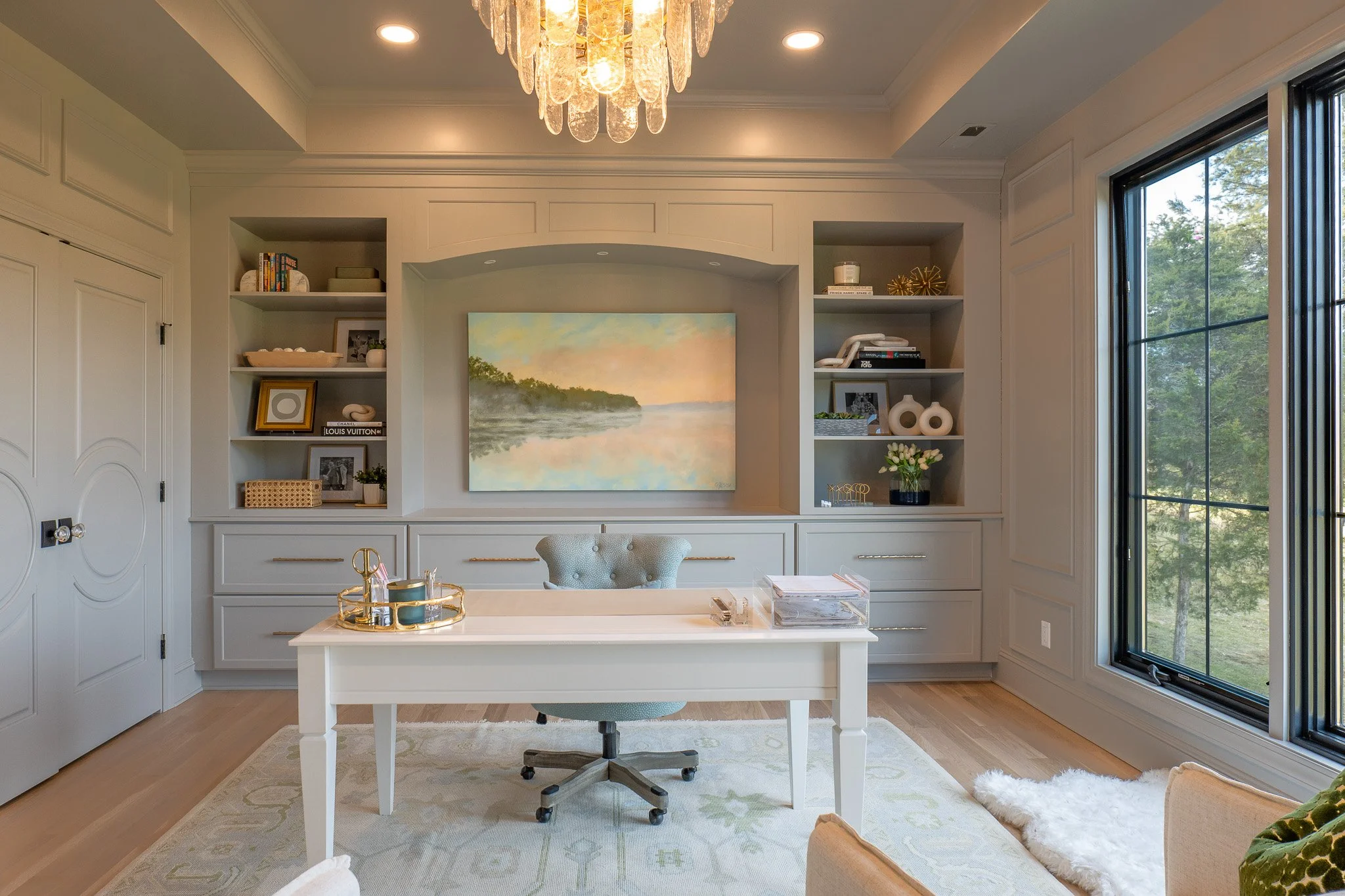 A bright home office with white walls, a white desk, an upholstered office chair, and built-in shelving filled with decorative items. Large window with greenery outside and a modern chandelier hanging from the ceiling.