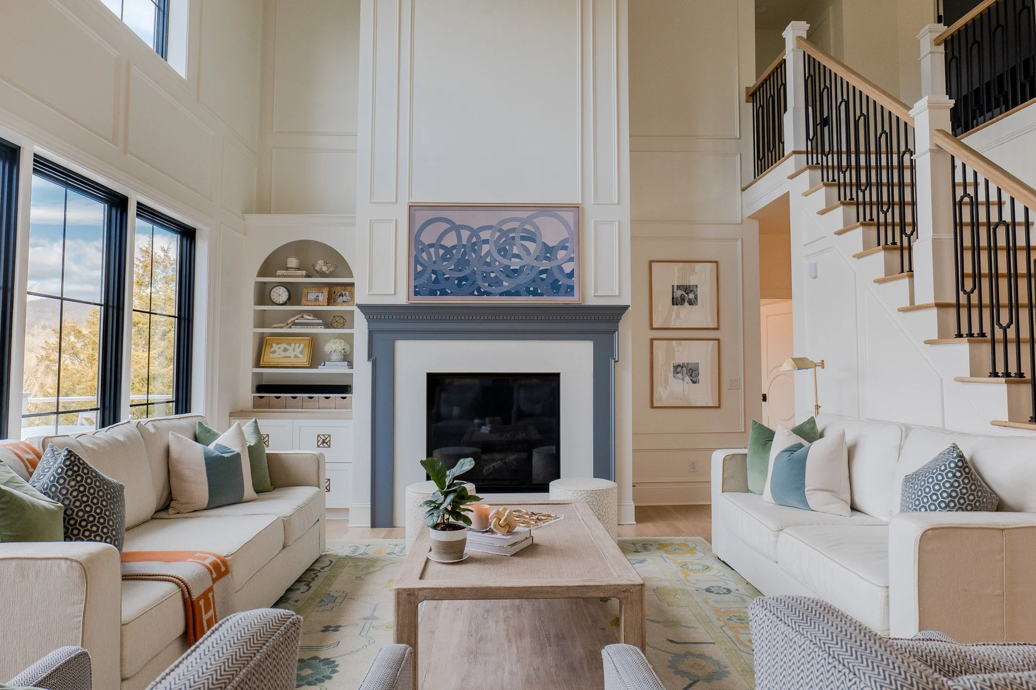 Living room with cream sofas, colorful pillows, a wooden coffee table, a white fireplace with a blue frame, large windows, and a staircase with black iron railing.