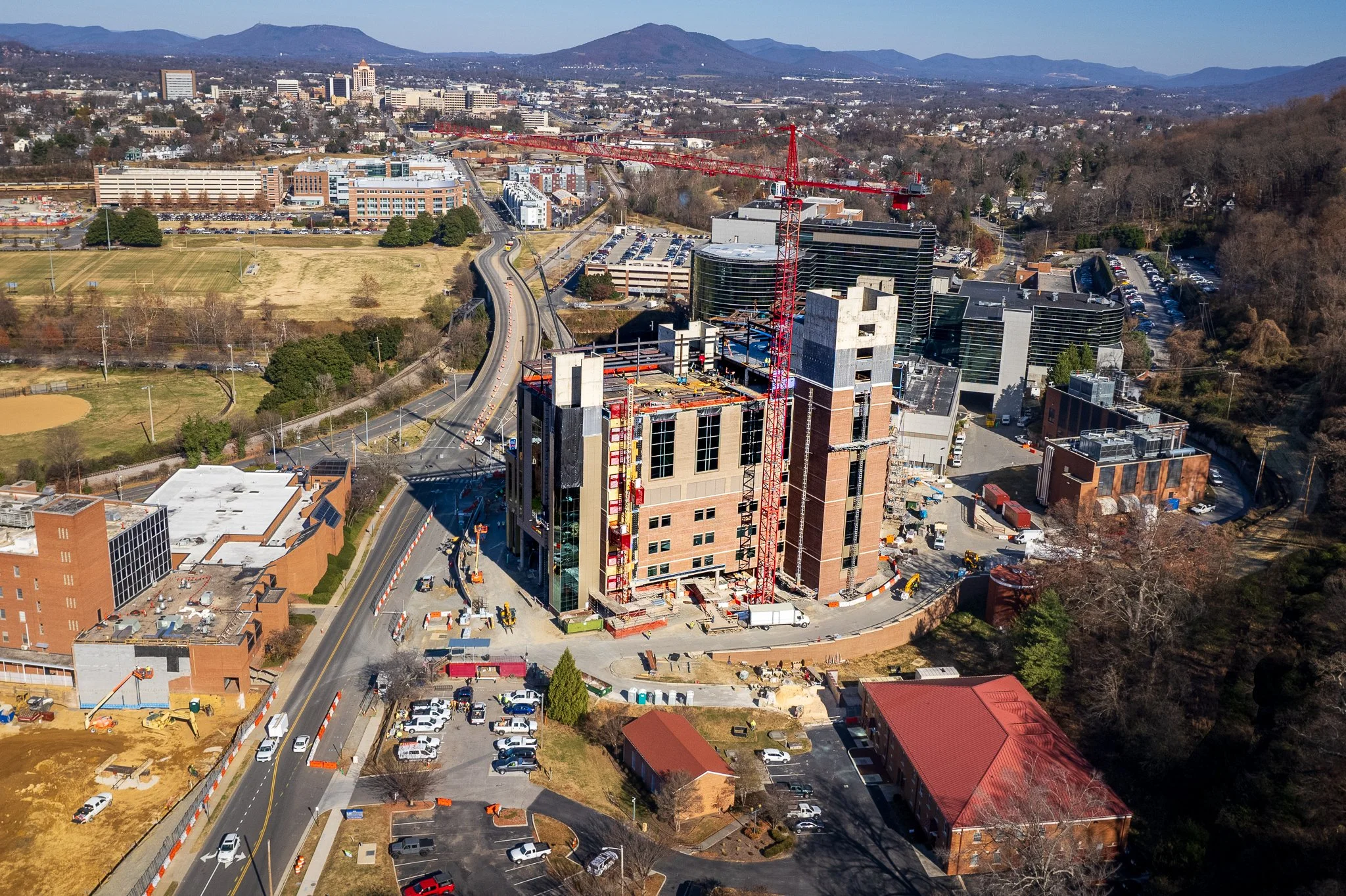 Aerial view of a cityscape with a construction site featuring a tall building under construction and a red crane, surrounded by roads, parking lots, and a mix of modern and older buildings, with mountains in the background.
