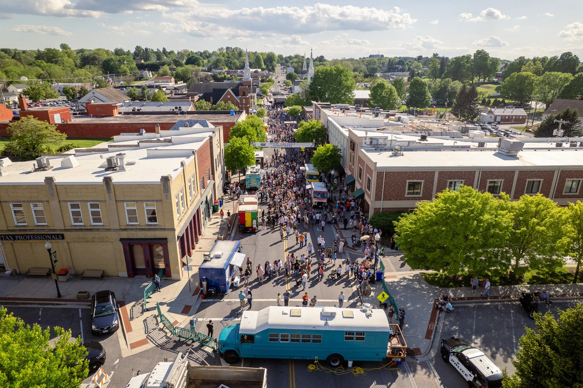 Aerial view of a busy downtown street filled with people, food trucks, and vendors during an outdoor event, lined with brick buildings and green trees, with a church and a partially cloudy sky in the background.
