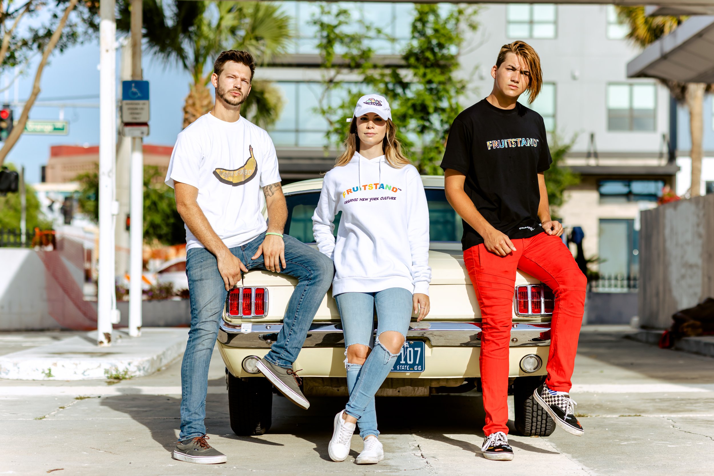 Three young people are posing in front of a vintage yellow car in an urban setting, with palm trees and buildings in the background. The person on the left is wearing a white t-shirt, blue jeans, and sneakers. The person in the middle is wearing a wh
