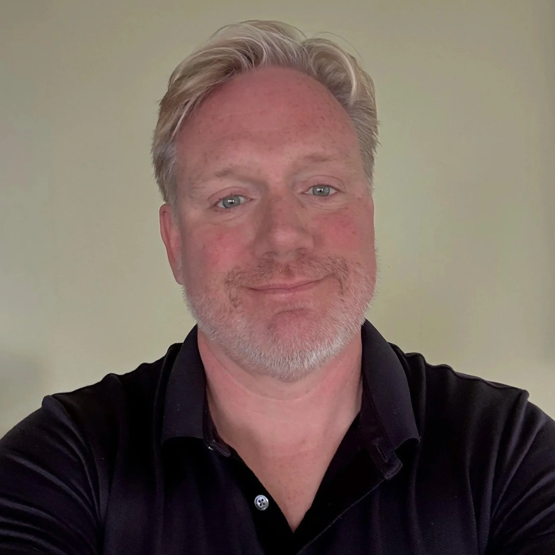 A bearded man with short-cropped hair and blue eyes smiles warmly directly at the camera.