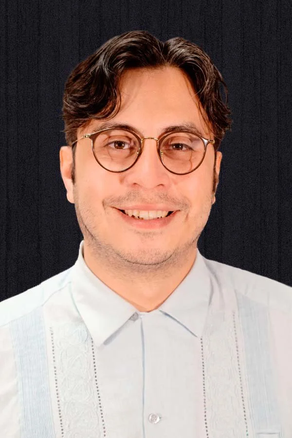 A young man with short brown hair and round-rimmed glasses smiles warmly at the camera.