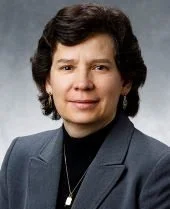 A woman smiles with joy at the camera. Her face is framed by short-cropped brown hair, she is wearing a grey blazer over a black shirt.