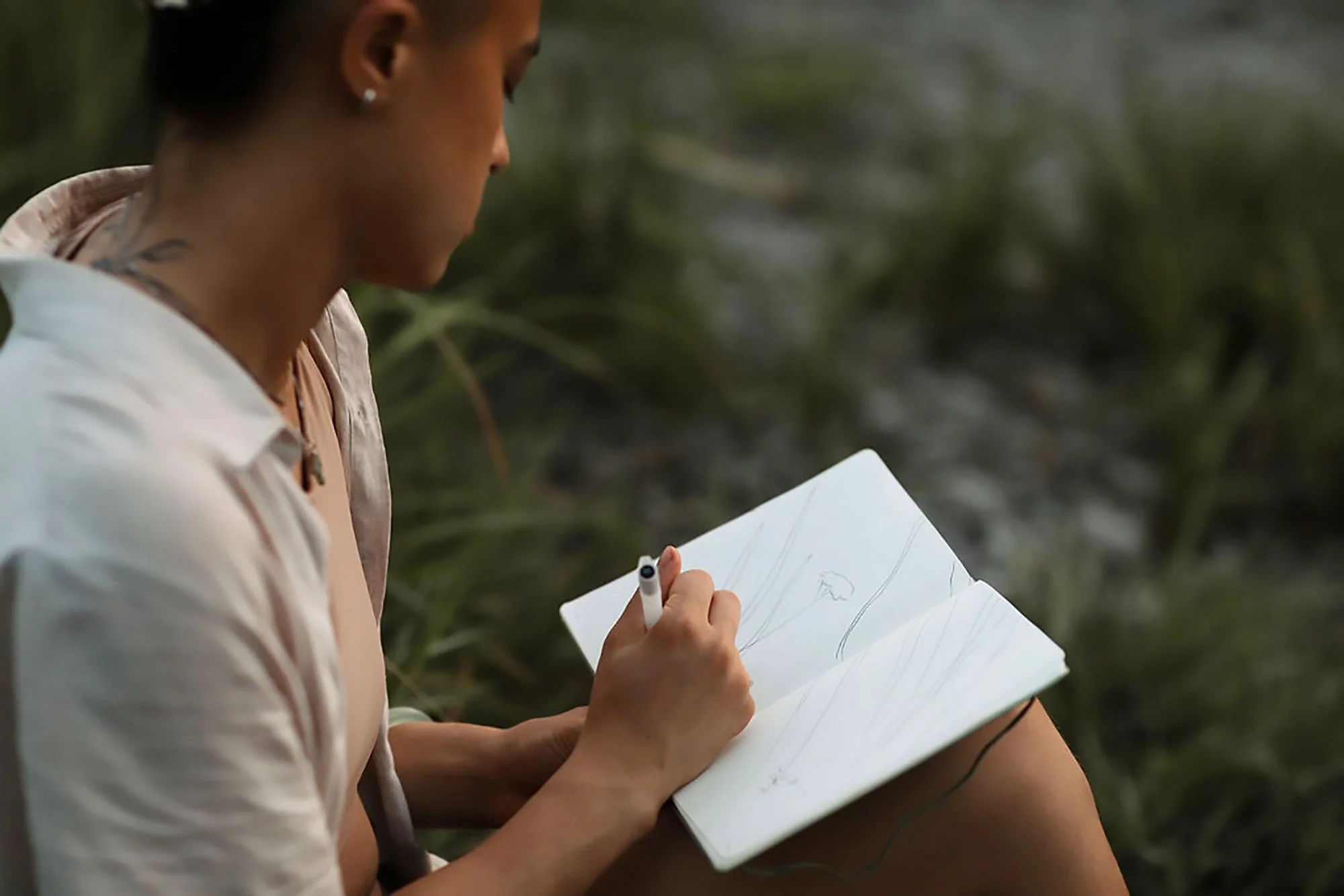 A woman sketching in a notebook outdoors with greenery in the background.