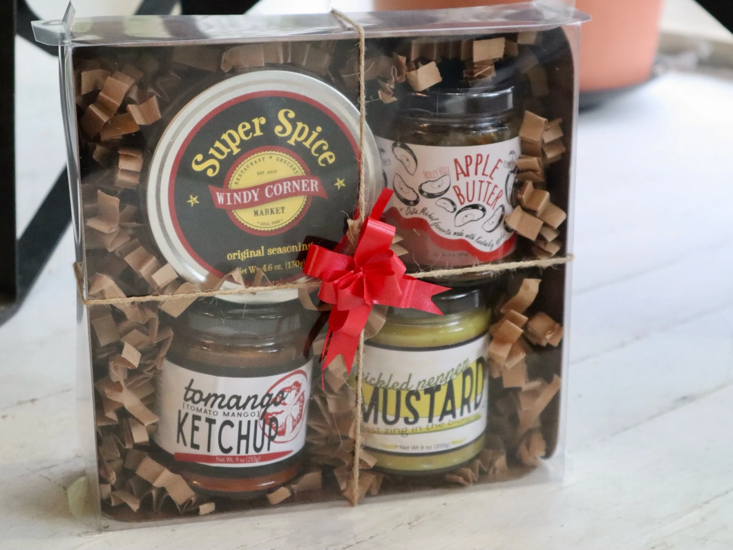 Gift set with four jars of condiments and seasonings inside a clear plastic box with brown shredded paper filler and a red ribbon across the front.