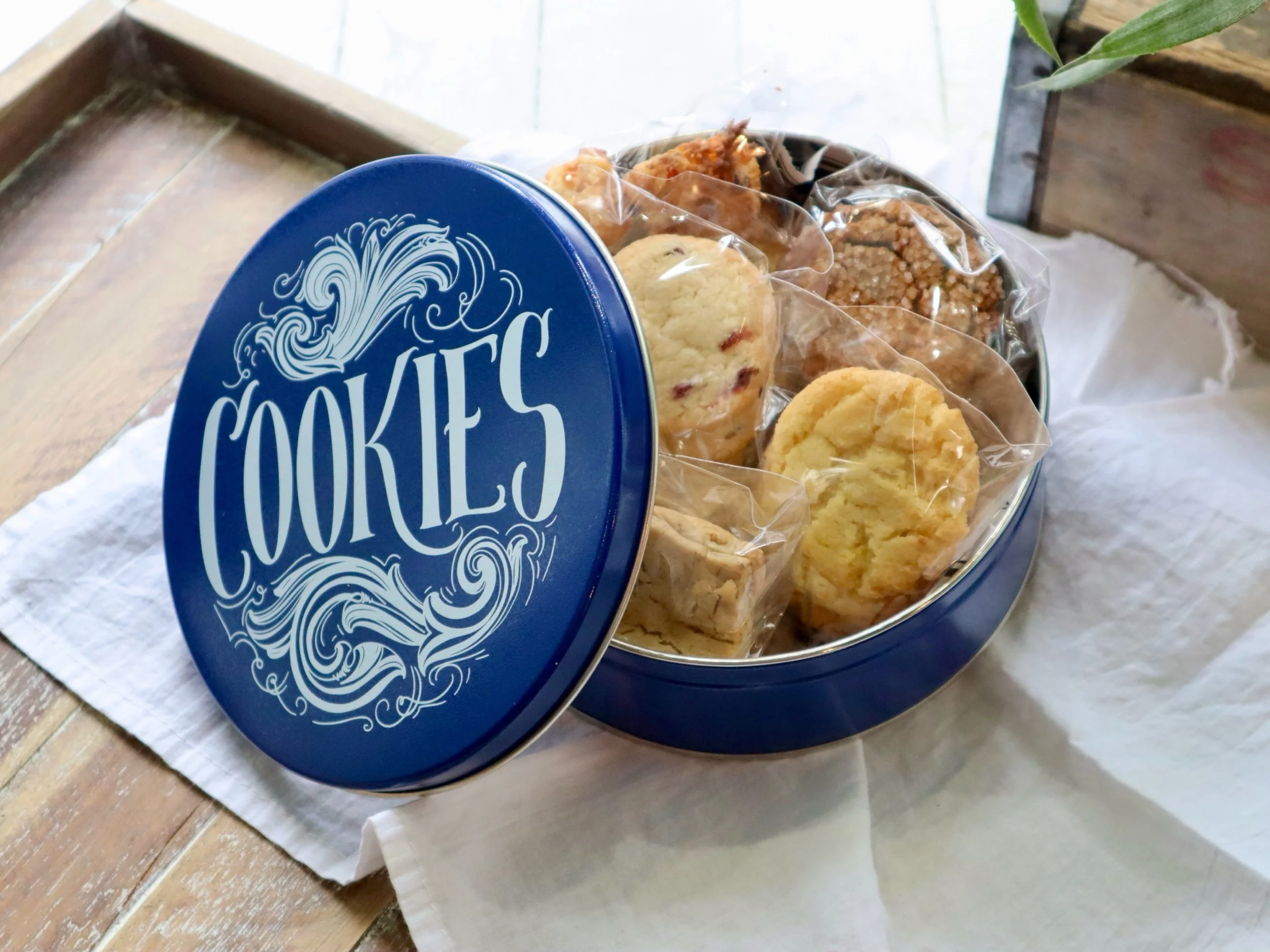 Round blue tin labeled 'Cookies' with a variety of individually wrapped cookies inside, resting on a white cloth on a wooden table.