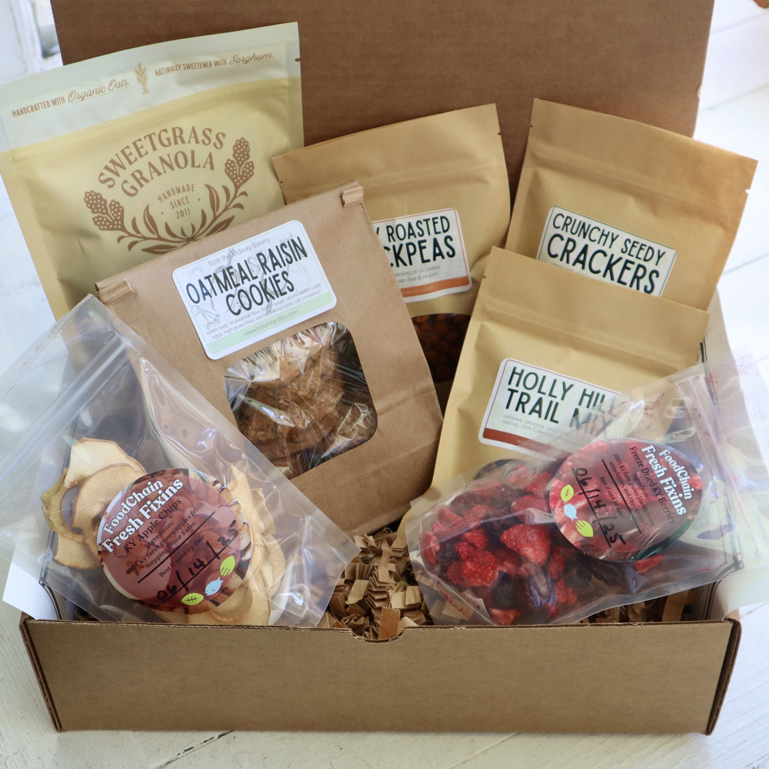Box containing assorted snacks including packets of granola, baked chickpeas, crackers, oatmeal raisin cookies, dried apples, and dried berries.