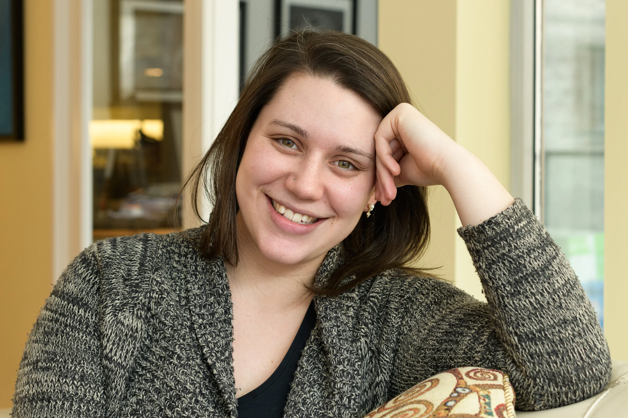 A young woman with brown hair and green eyes smiling and resting her head on her hand, sitting indoors near windows.