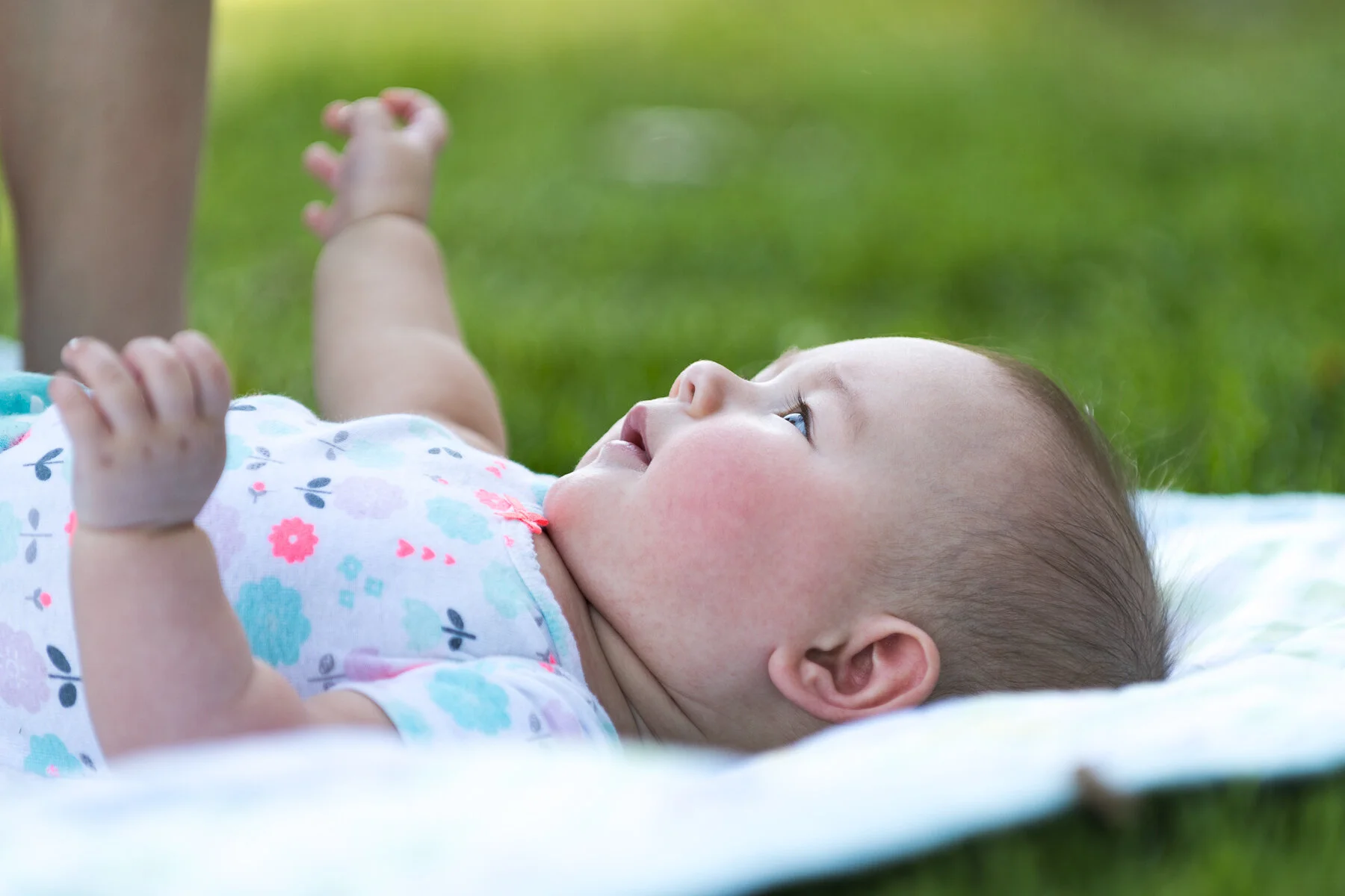 A baby lying on a white blanket outdoors on green grass, looking up and smiling.