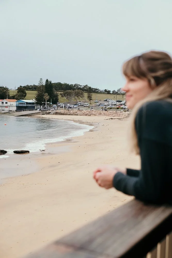 A woman standing on a wooden railing overlooking a sandy beach with a few rocks and gentle waves, with a parking lot, campus buildings, and green hills in the background.